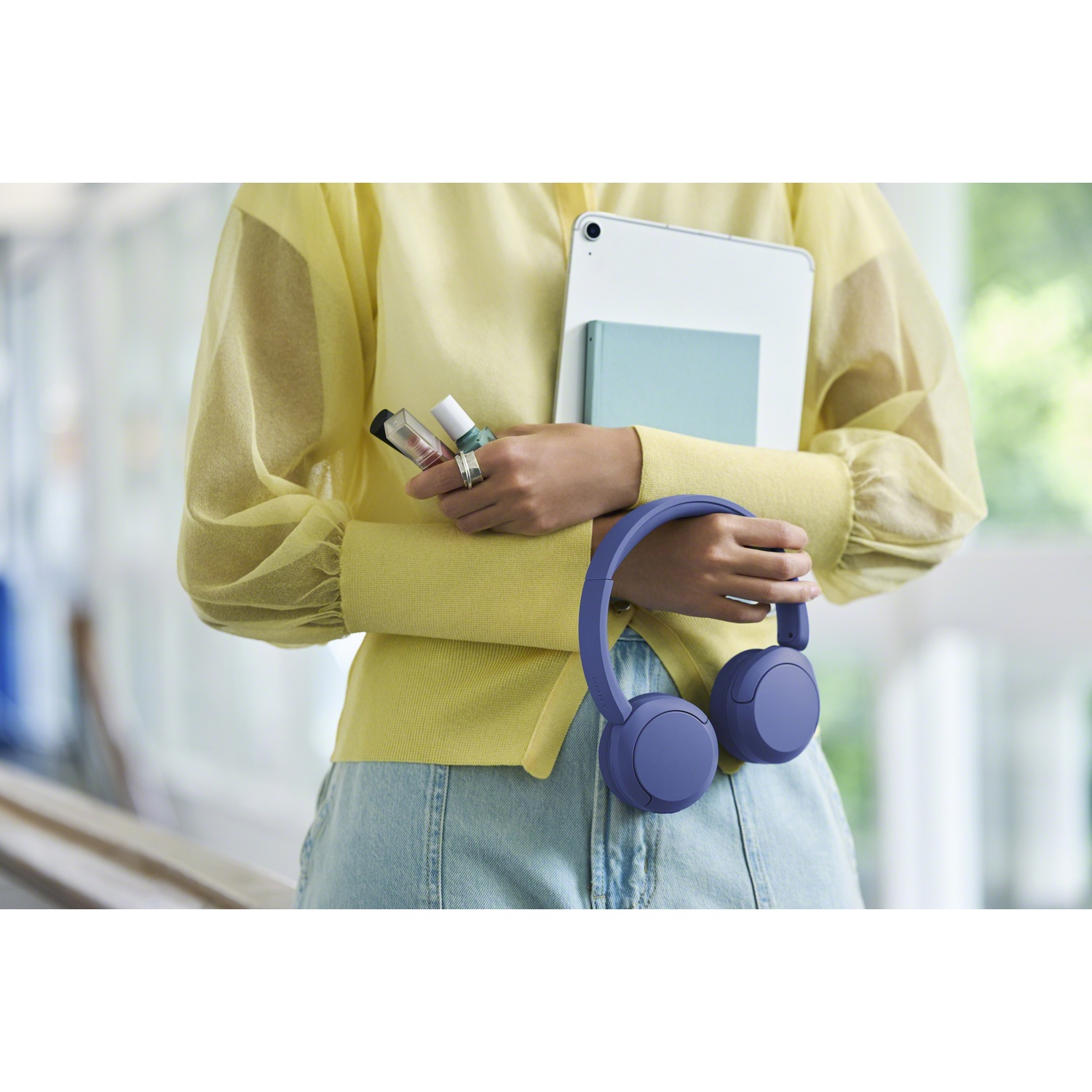Une personne en chemisier jaune tient une tablette, du rouge à lèvres et un casque violet.