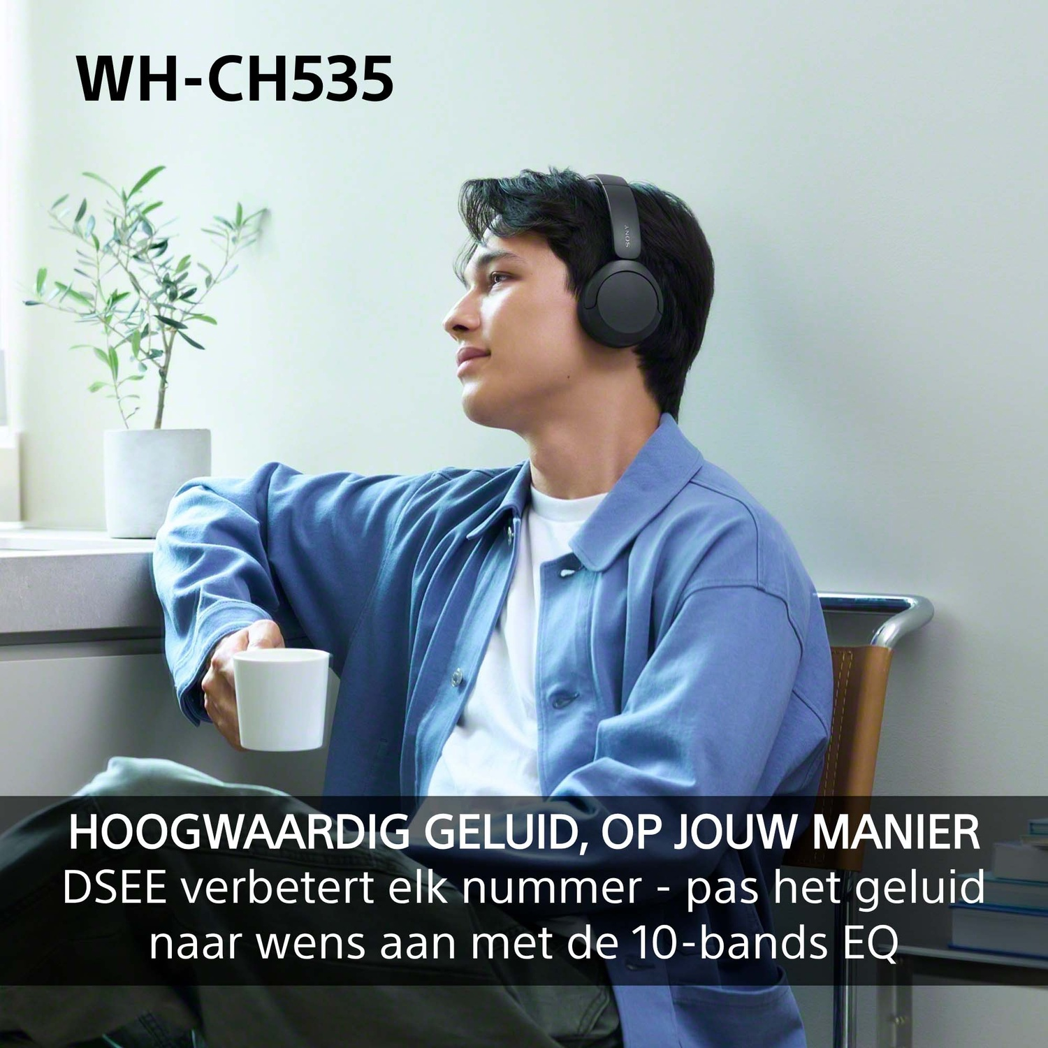 Een jonge man in een blauwe jas en koptelefoon houdt een mok vast in een kamer met plant.