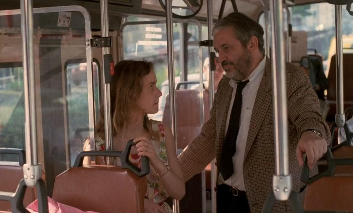 Ein Mann in karierter Jacke und Krawatte steht in einem Bus und spricht mit einer jungen Frau.