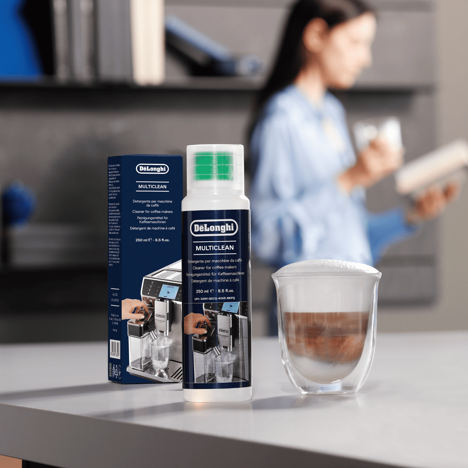 Limpiador DeLonghi Multiclean y taza de café con espuma en una mesa, mujer al fondo.