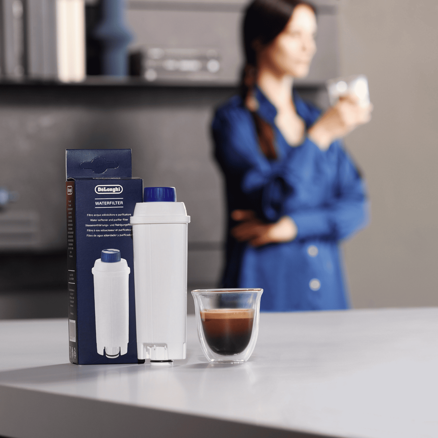 Un filtro de agua DeLonghi con su caja y una taza de espresso sobre una mesa. Una mujer de pie al fondo.