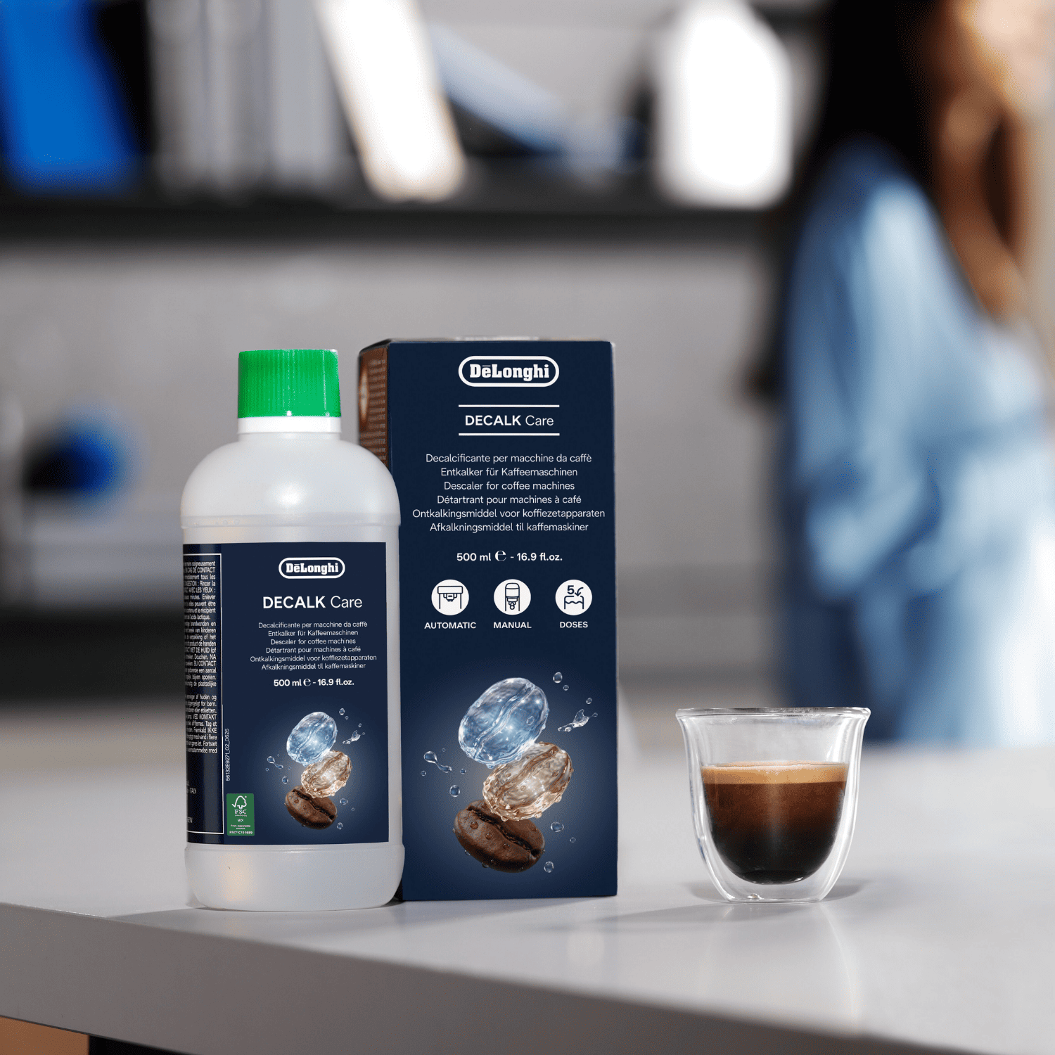 Una botella y caja de líquido descalcificante DeLonghi en un mostrador blanco junto a un espresso.