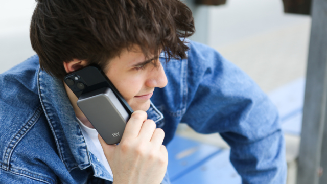Un jeune homme aux cheveux bruns et en veste en jean téléphone avec un smartphone muni d'une batterie externe grise.