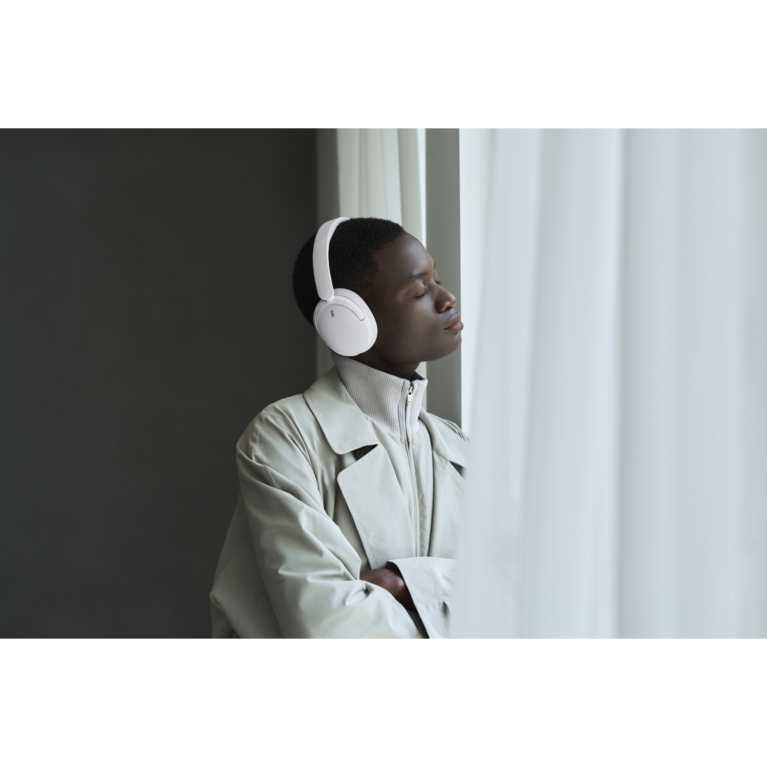 Un homme noir portant un casque blanc et un manteau beige se tient près d'une fenêtre avec des rideaux blancs.