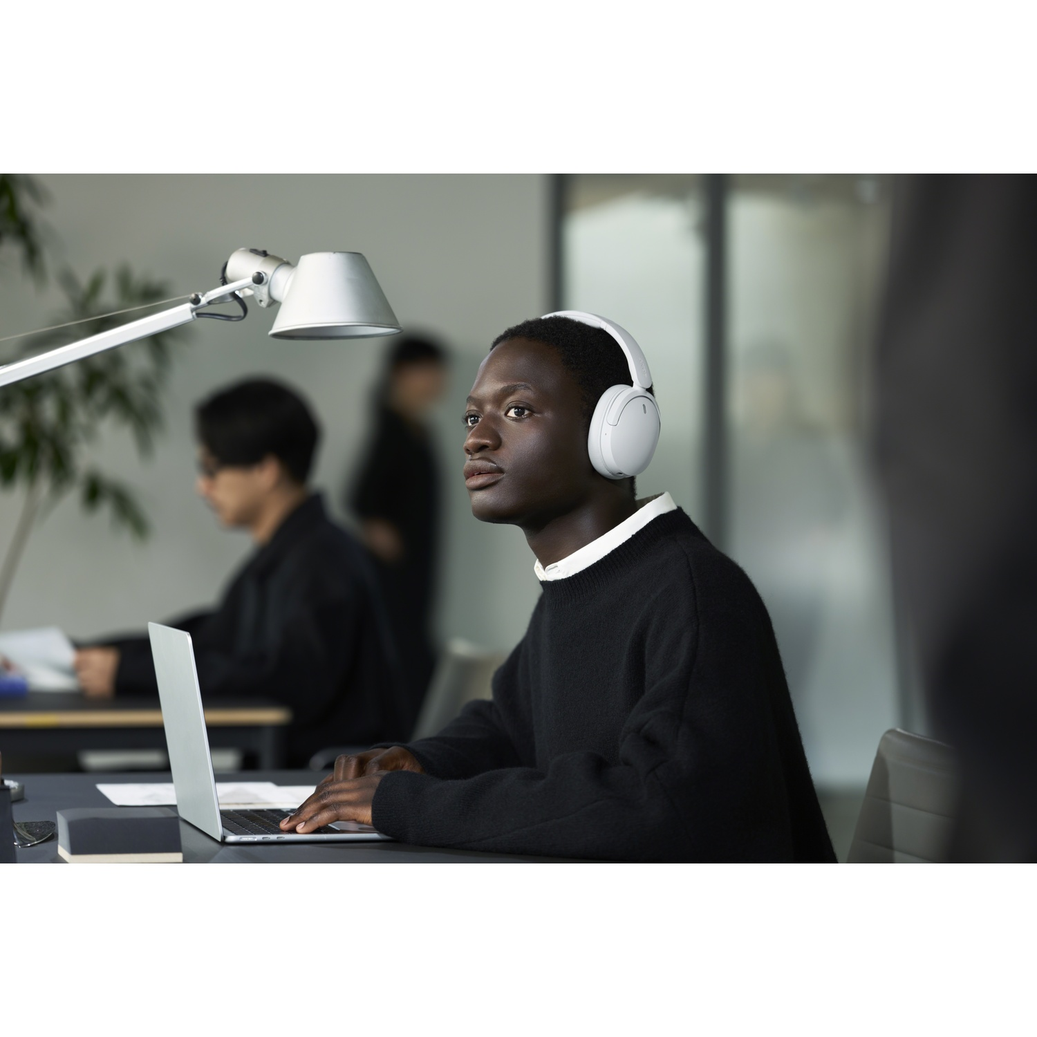 Un jeune homme portant un casque blanc tape sur un ordinateur portable dans un bureau, d'autres travaillant en arrière-plan.