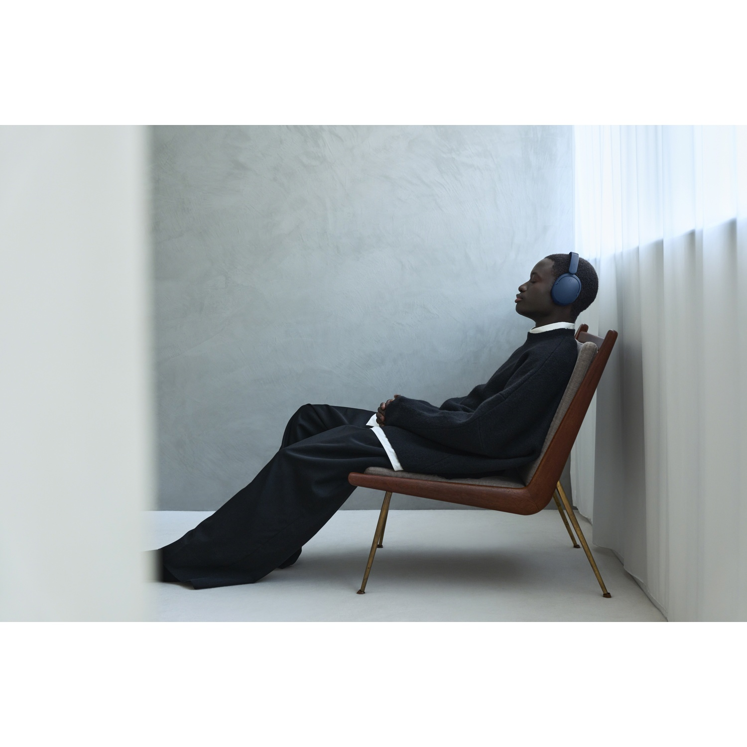 Un homme les yeux fermés et des écouteurs bleus est assis dans une chaise en bois contre un mur gris.