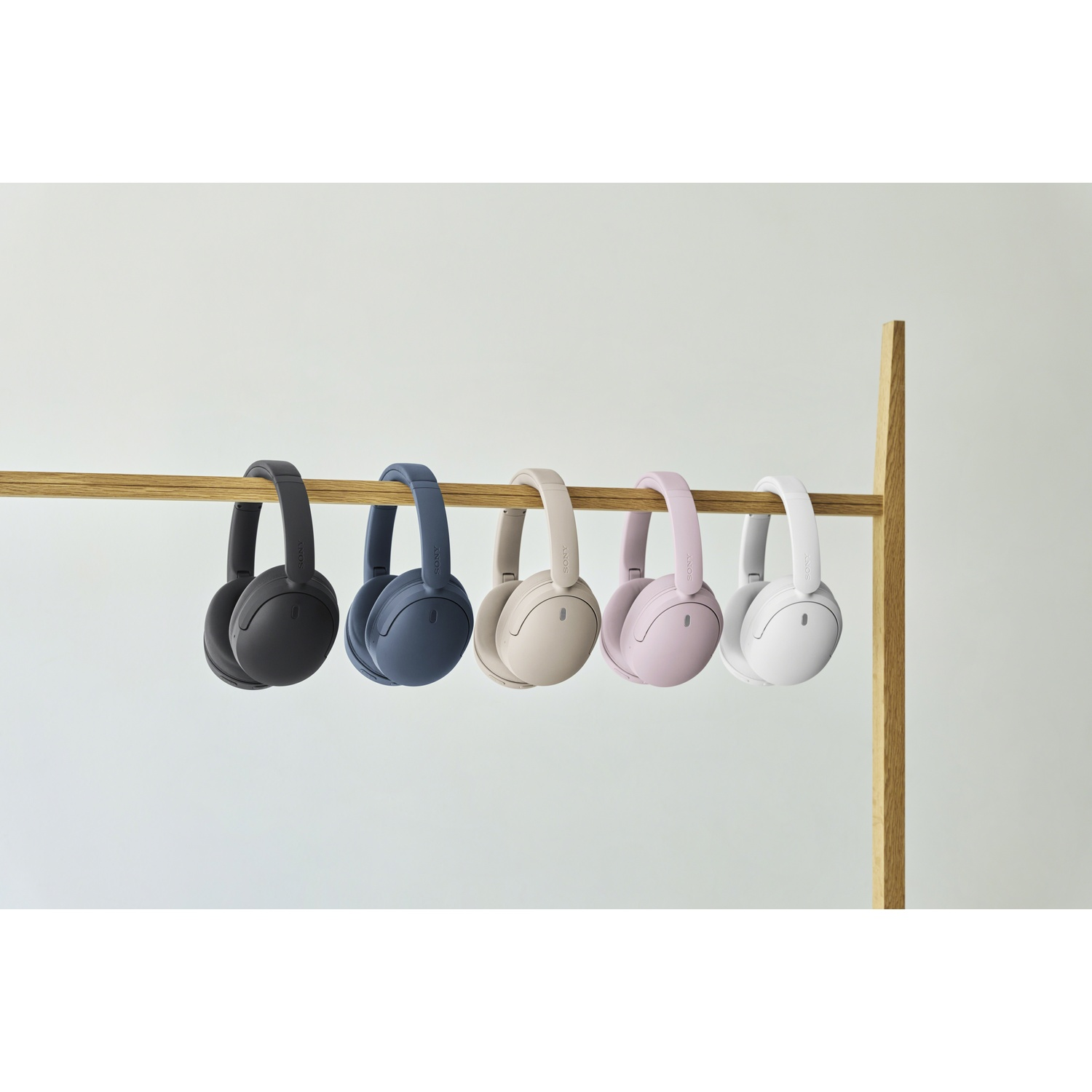 Cinq paires de casques sans fil noirs, bleus, beiges, roses et blancs pendent à une barre en bois sur fond clair.