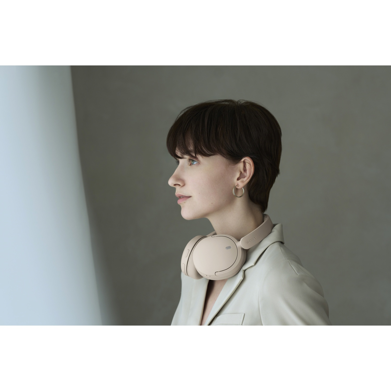 Une femme aux cheveux courts et au casque beige autour du cou se tient dans une pièce contre un mur gris.