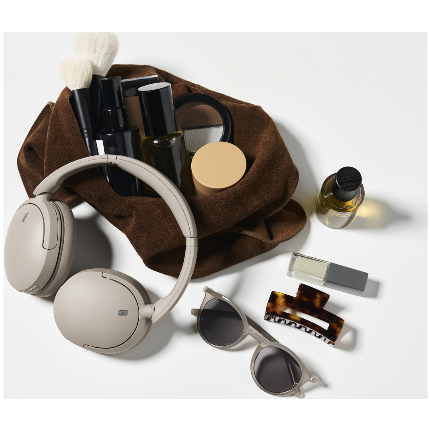 Une pochette en daim marron avec des cosmétiques, des pinceaux, un casque beige, des lunettes de soleil et du parfum.