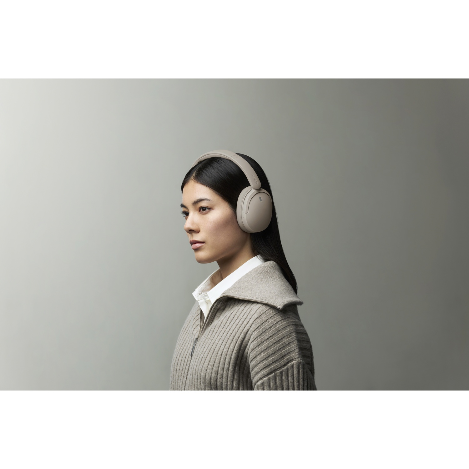 Une femme aux cheveux noirs, chemise blanche et pull beige porte un casque beige sur fond gris.