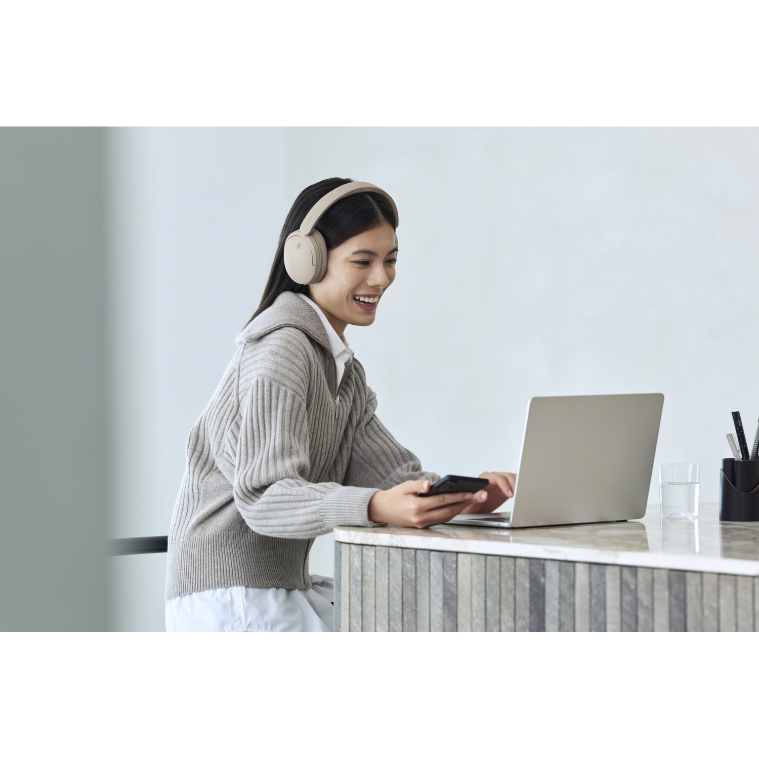 Une femme souriante portant un casque est assise à table, tenant un smartphone et utilisant un ordinateur.