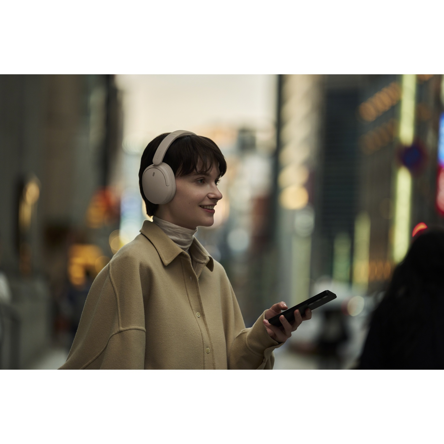 Une femme en veste beige portant un casque blanc tient un smartphone dans une rue de ville illuminée.