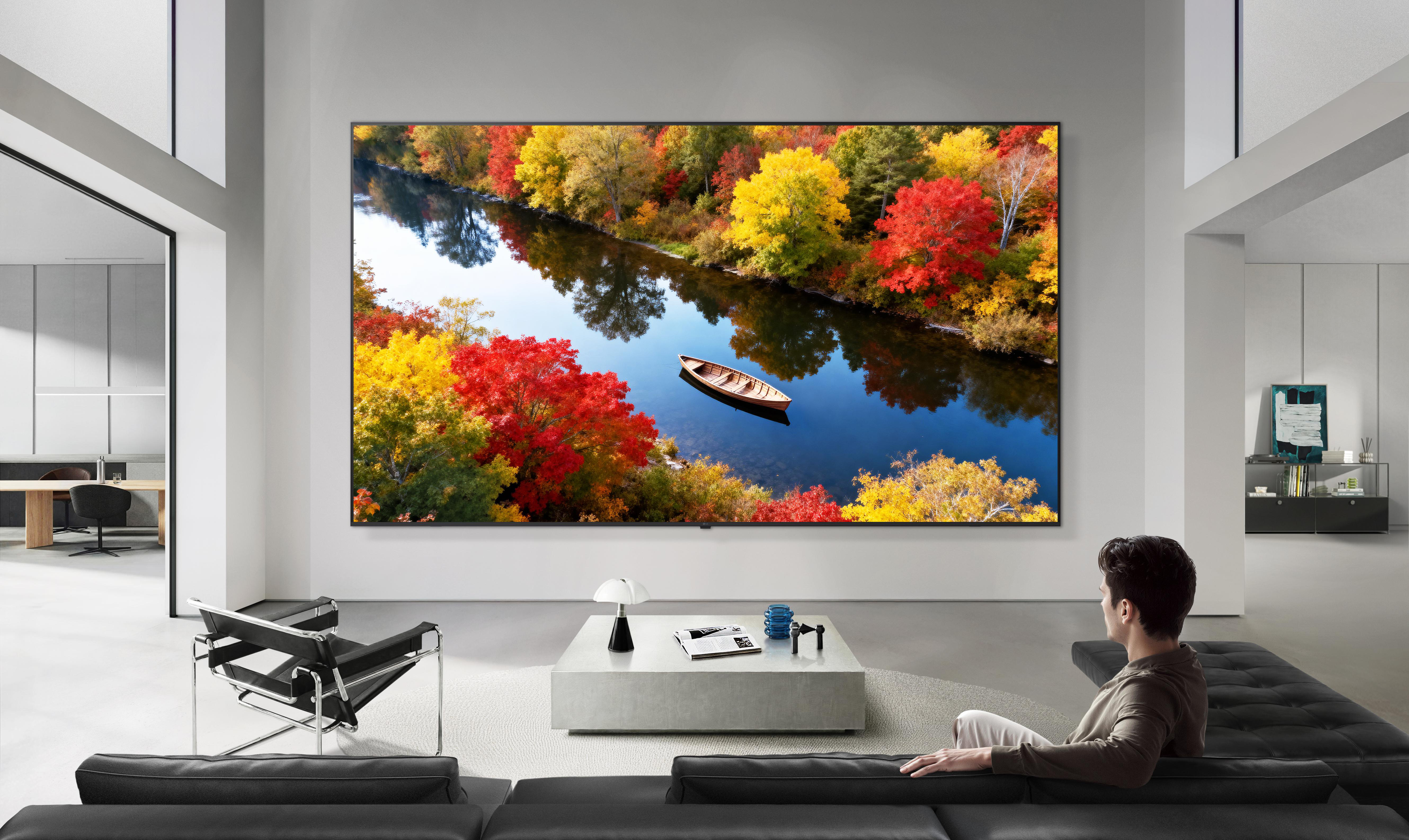 Ein Mann sitzt in einem modernen Wohnzimmer vor einem riesigen Bildschirm mit herbstlicher Flusslandschaft.