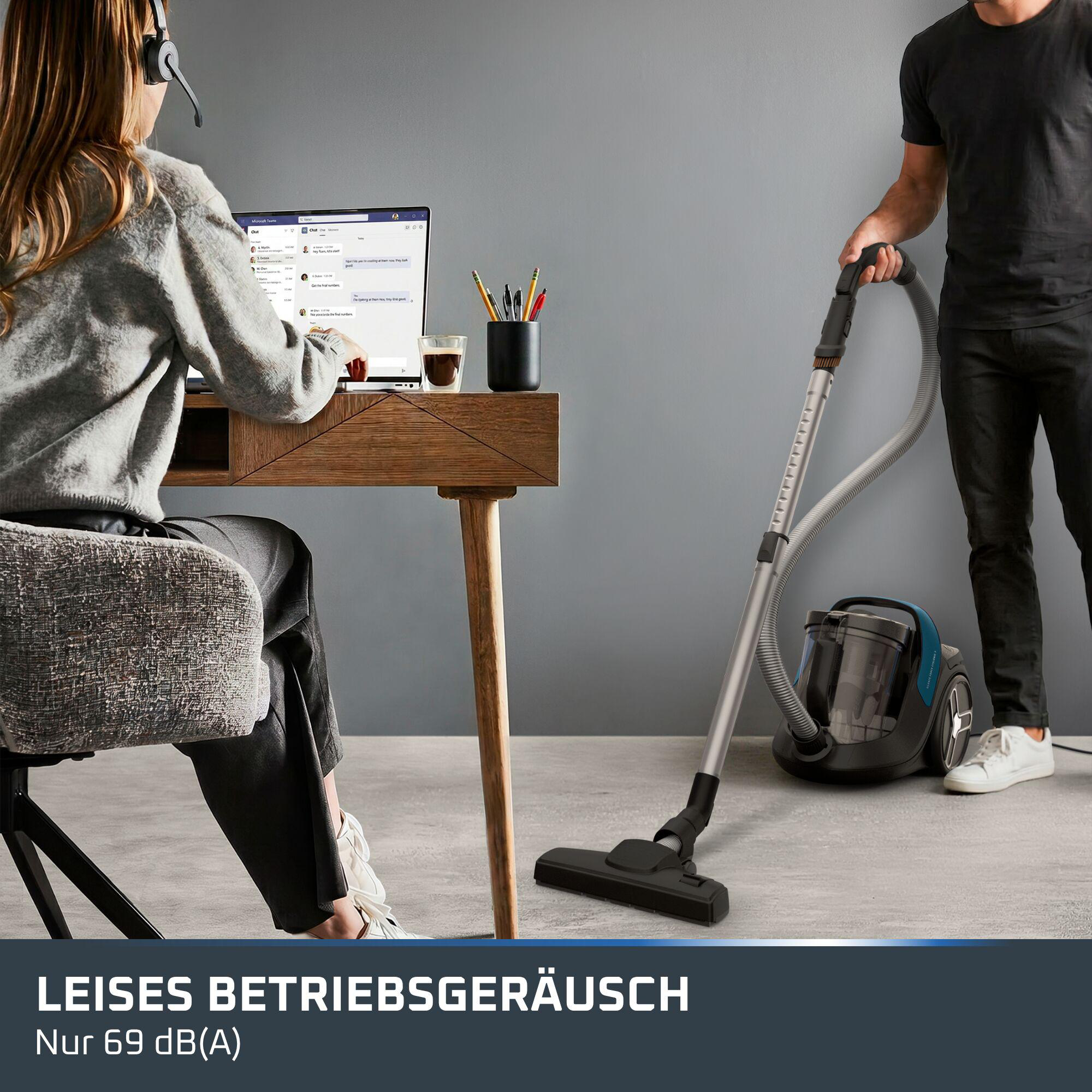 Eine Frau arbeitet am Laptop mit Headset, während ein Mann im Hintergrund mit einem Staubsauger saugt.