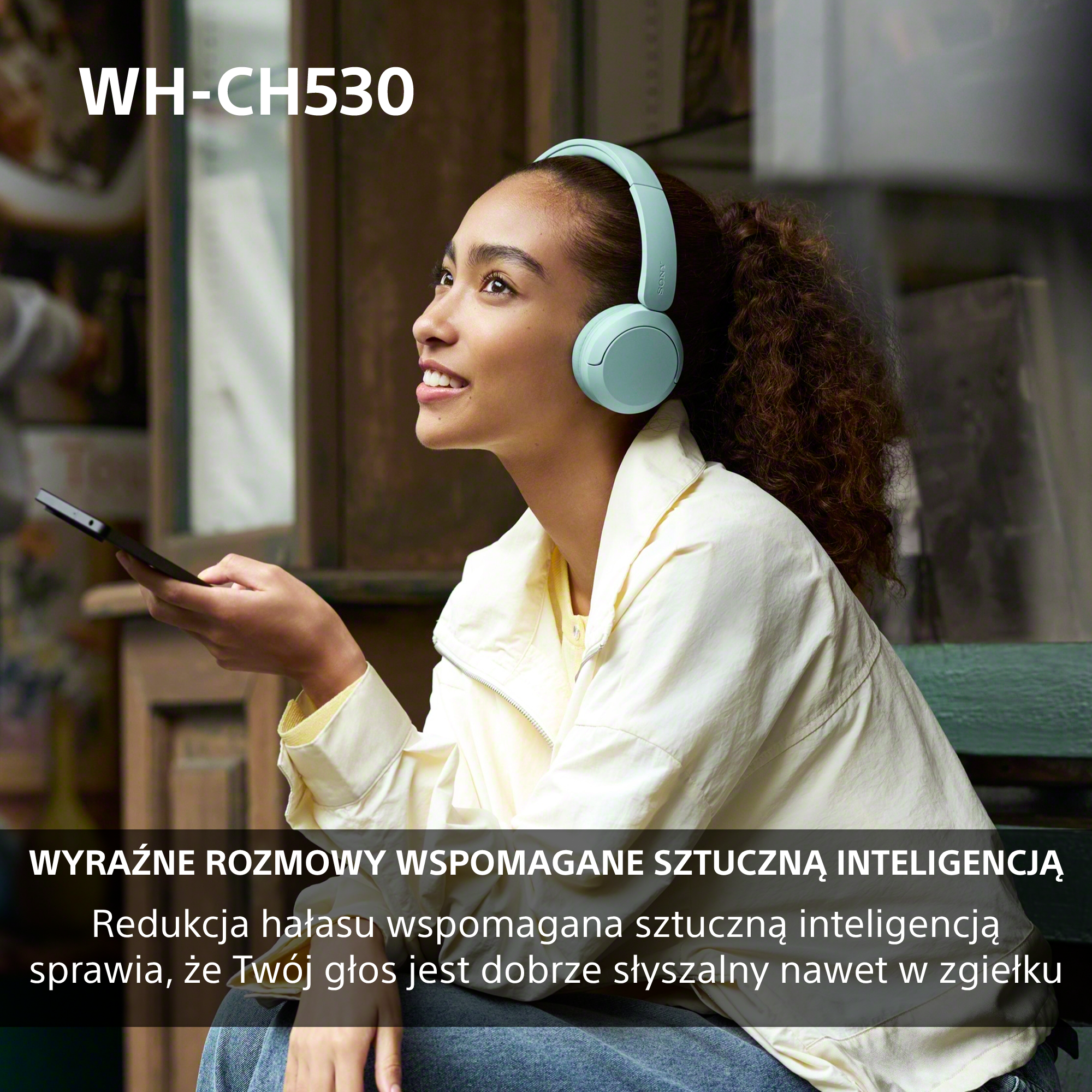 Kobieta w jasnozielonych słuchawkach i kremowej kurtce trzyma telefon, uśmiechając się.