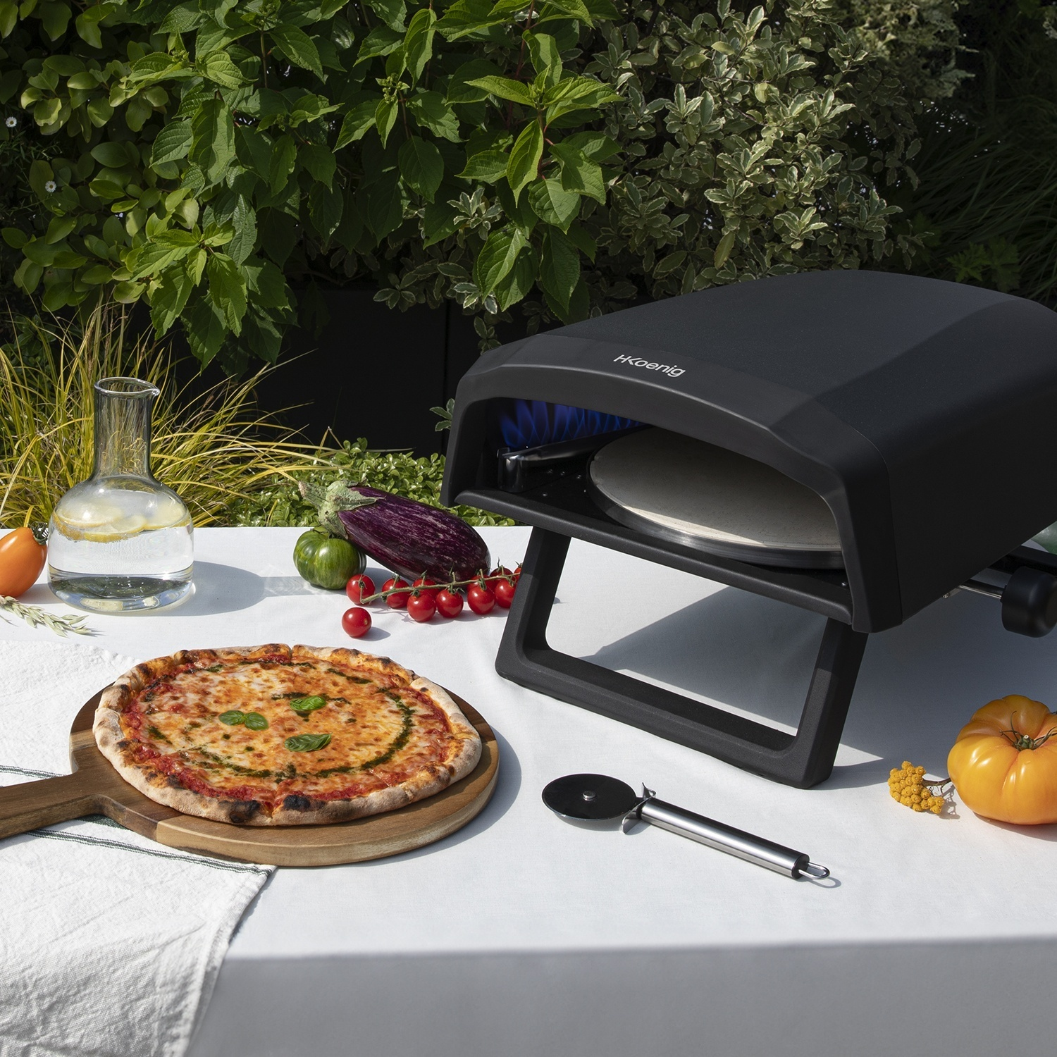 Un forno per pizza nero si trova all'aperto su un tavolo accanto a una pizza fresca, verdure e una brocca d'acqua.