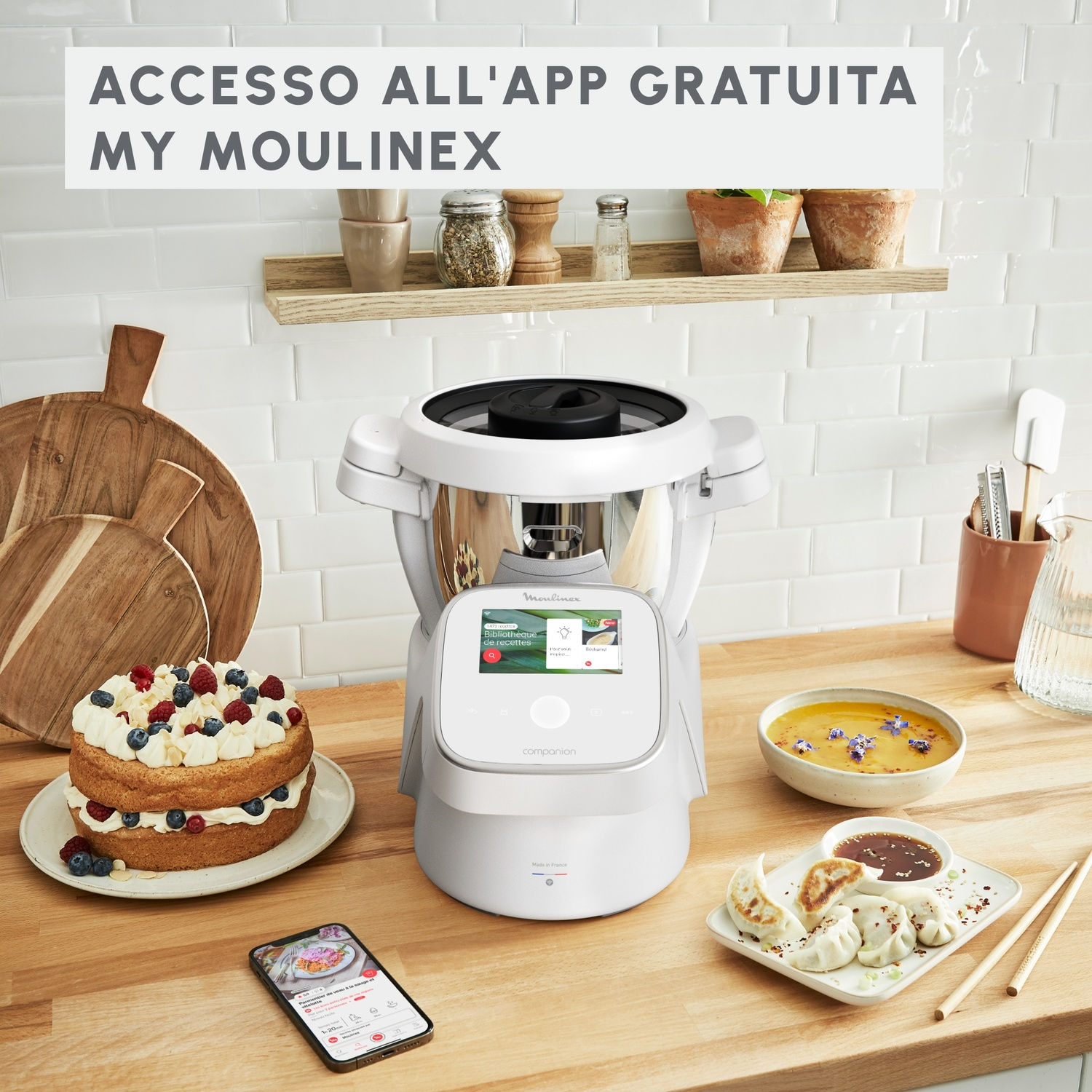 Un robot da cucina Moulinex bianco su un piano di lavoro con torta, zuppa, ravioli e smartphone.