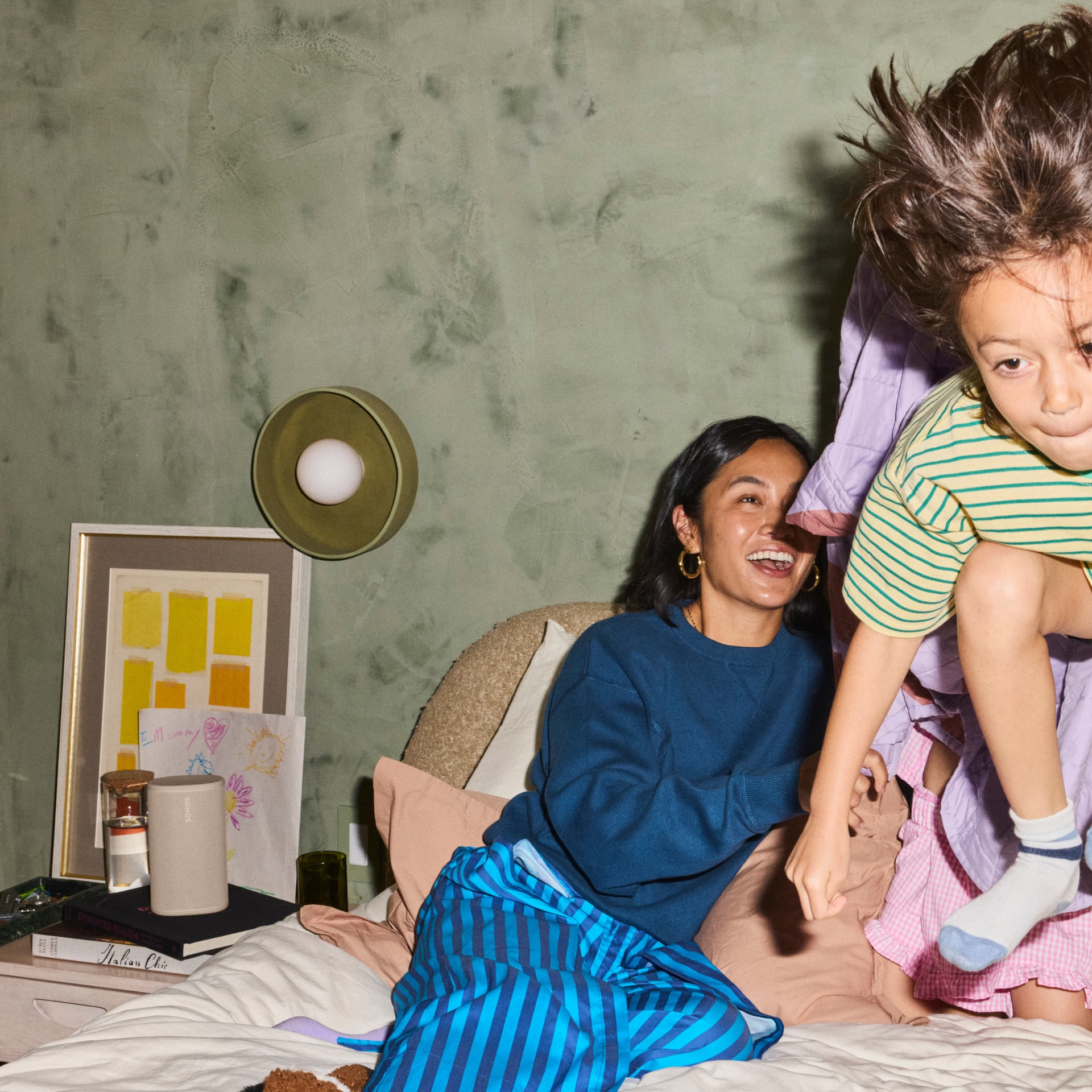 Une femme souriante est assise sur un lit pendant qu'un enfant saute joyeusement.