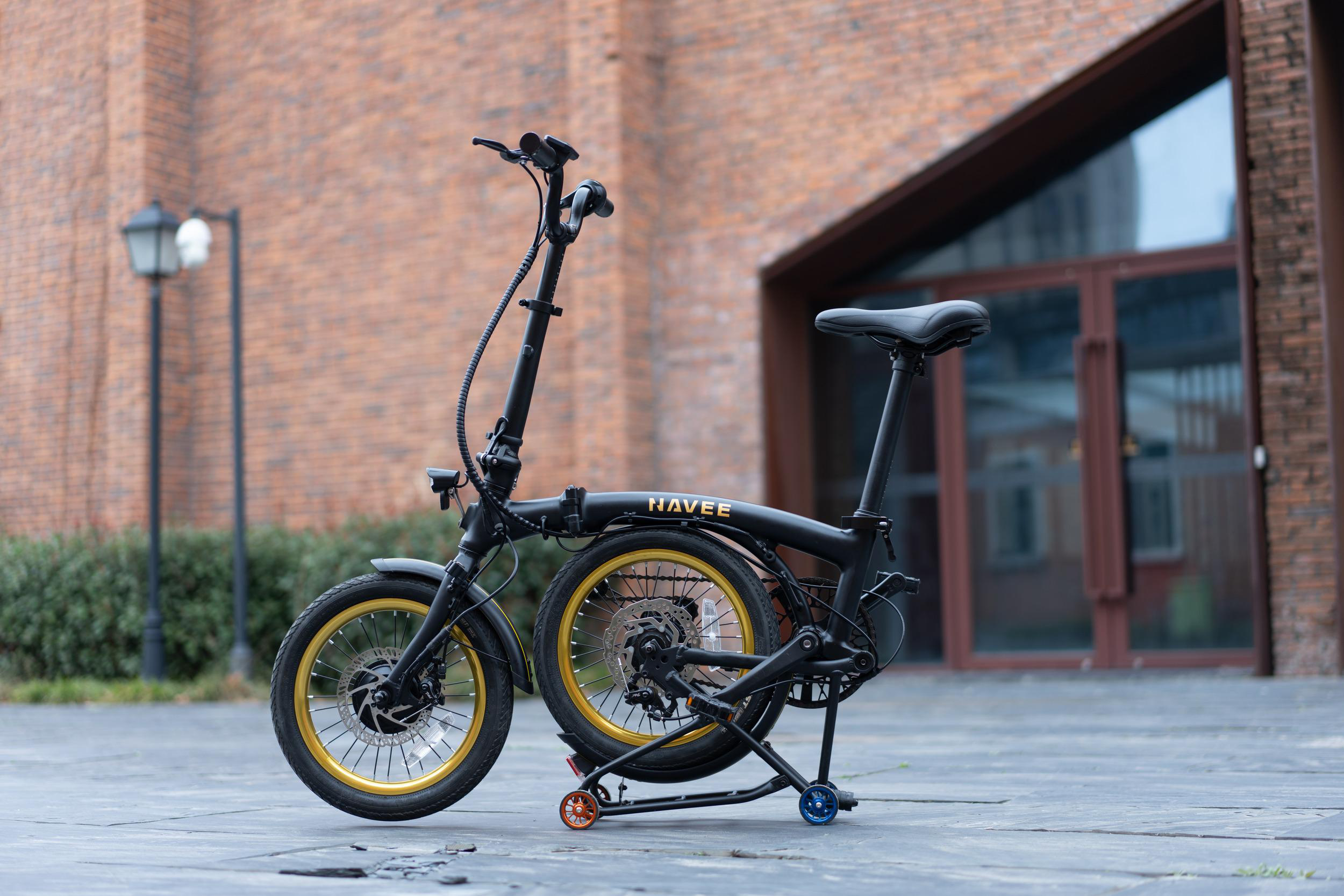 Ein schwarzes Faltfahrrad mit goldenen Felgen und dem NAVEE-Schriftzug steht auf grauen Fliesen vor einer Backsteinwand.