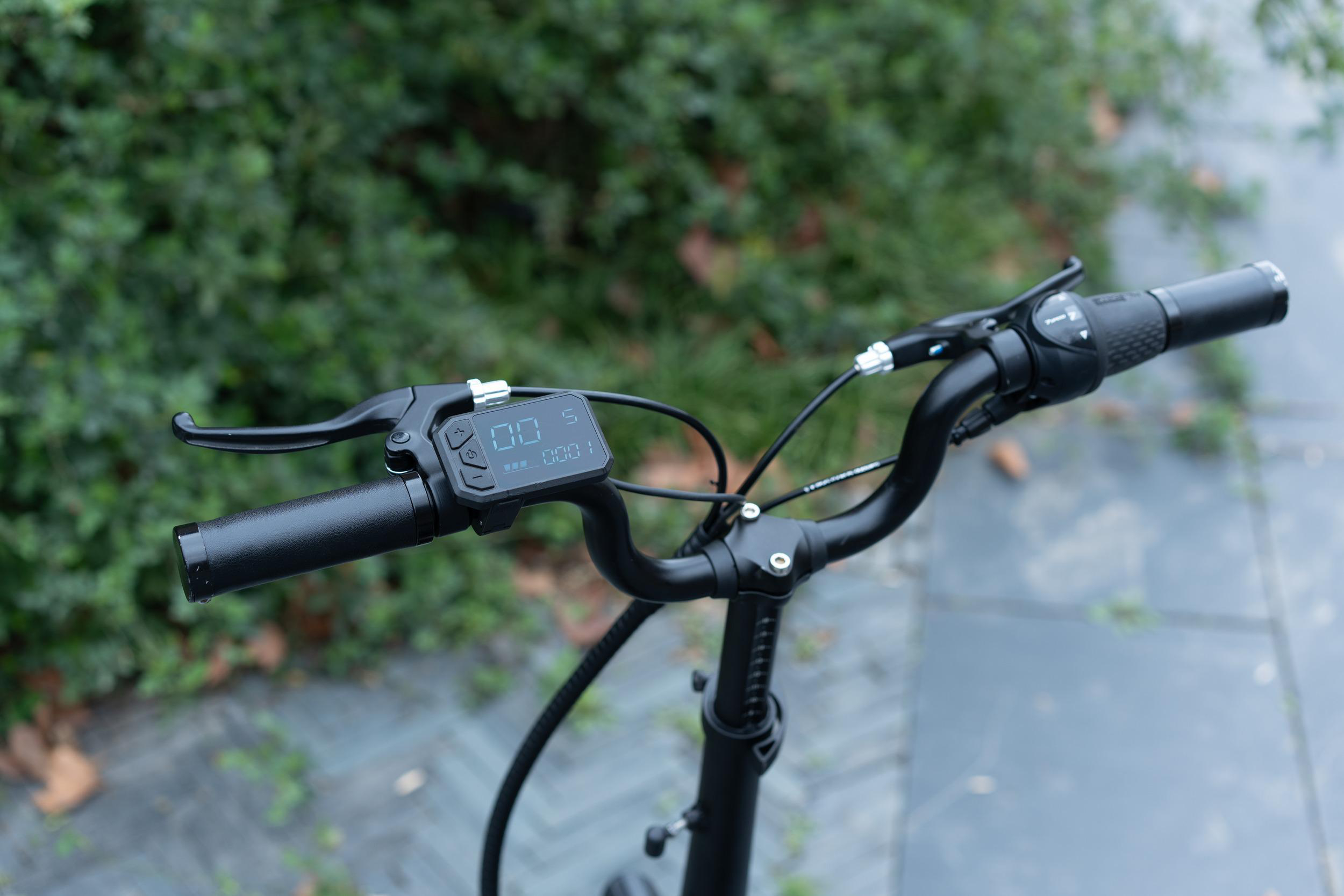 Schwarzer E-Bike-Lenker mit digitalem Display und Bremshebeln, draußen mit grünem Hintergrund.