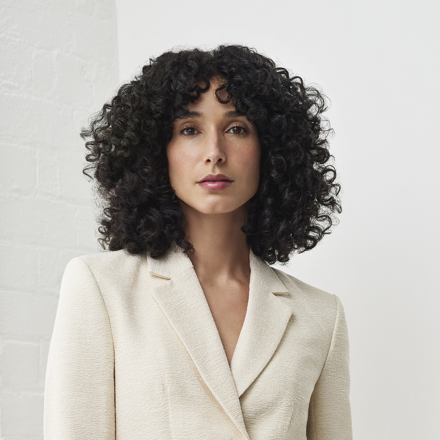 Ritratto di una donna con capelli scuri ricci e un blazer chiaro su sfondo bianco.