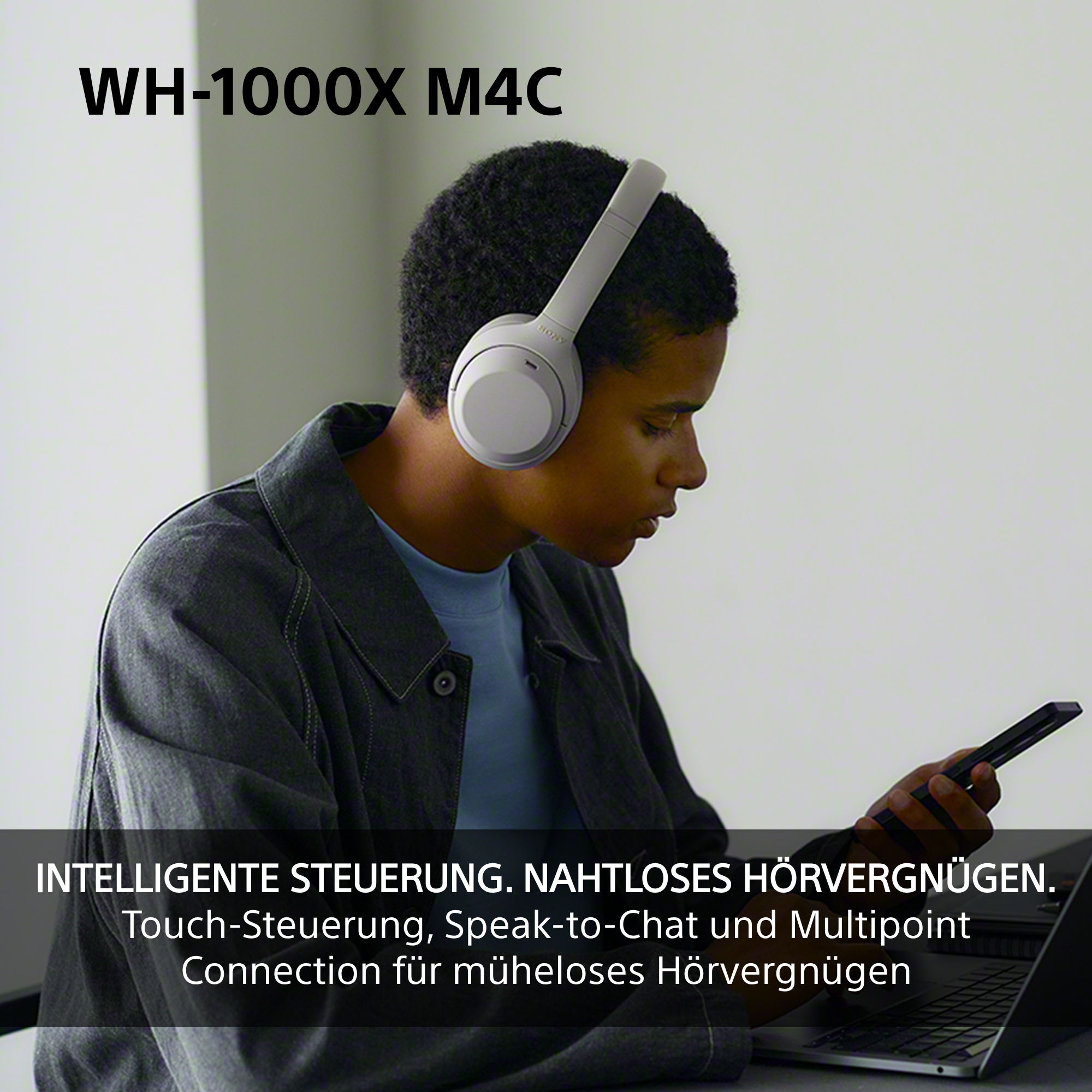 Mann mit grauen Kopfhörern und Smartphone sitzt vor einem Laptop.