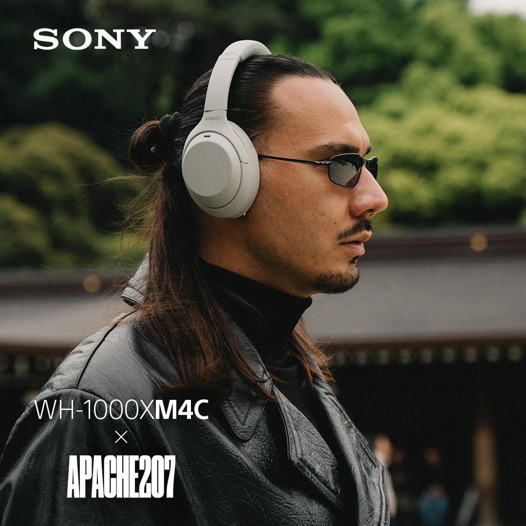 Ein Mann mit langem Haar, Sonnenbrille und Lederjacke trägt weiße Sony-Kopfhörer vor einer grünen Kulisse.