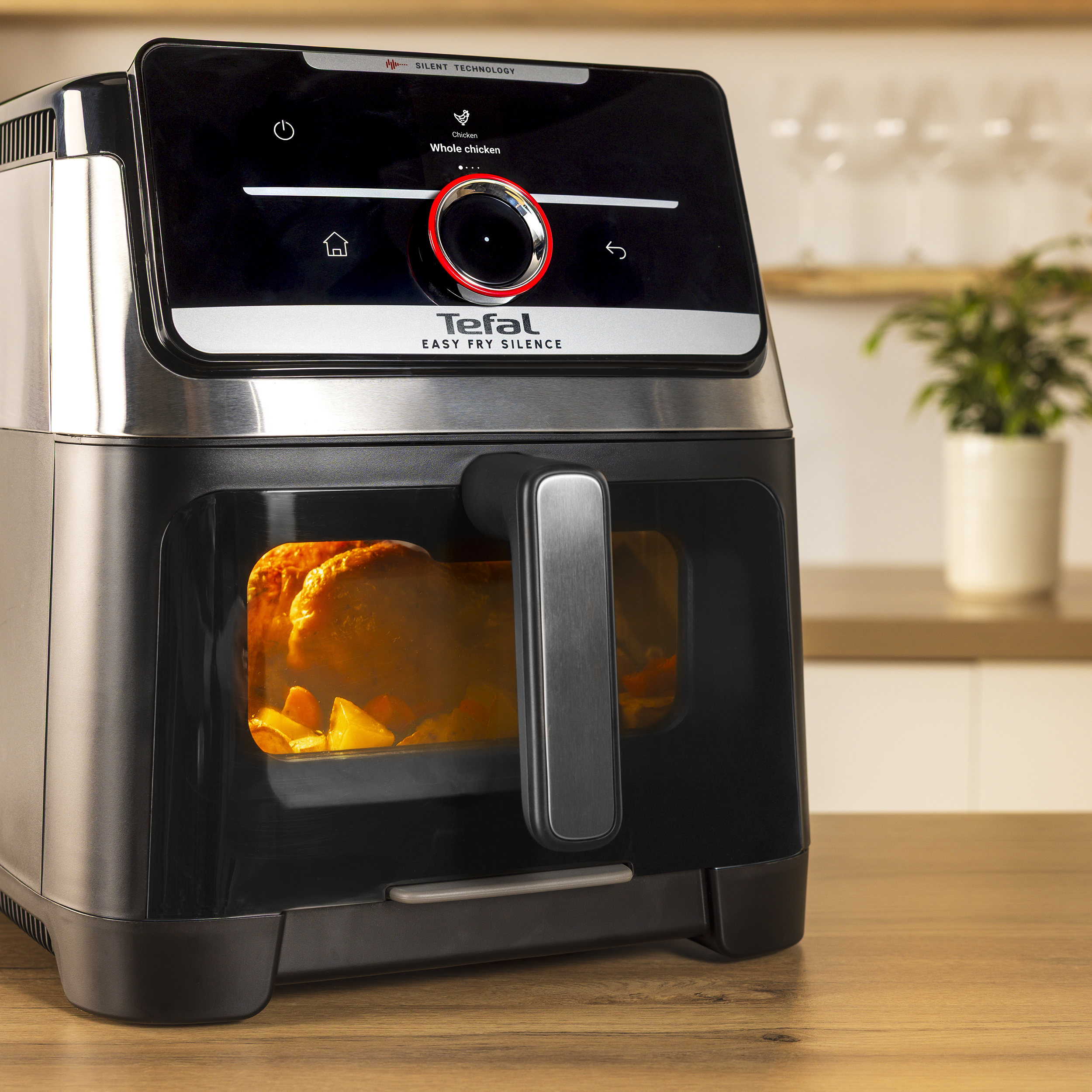 Fekete Tefal Easy Fry Silence légsütő fa pulton, világító ablakon át látható étellel.
