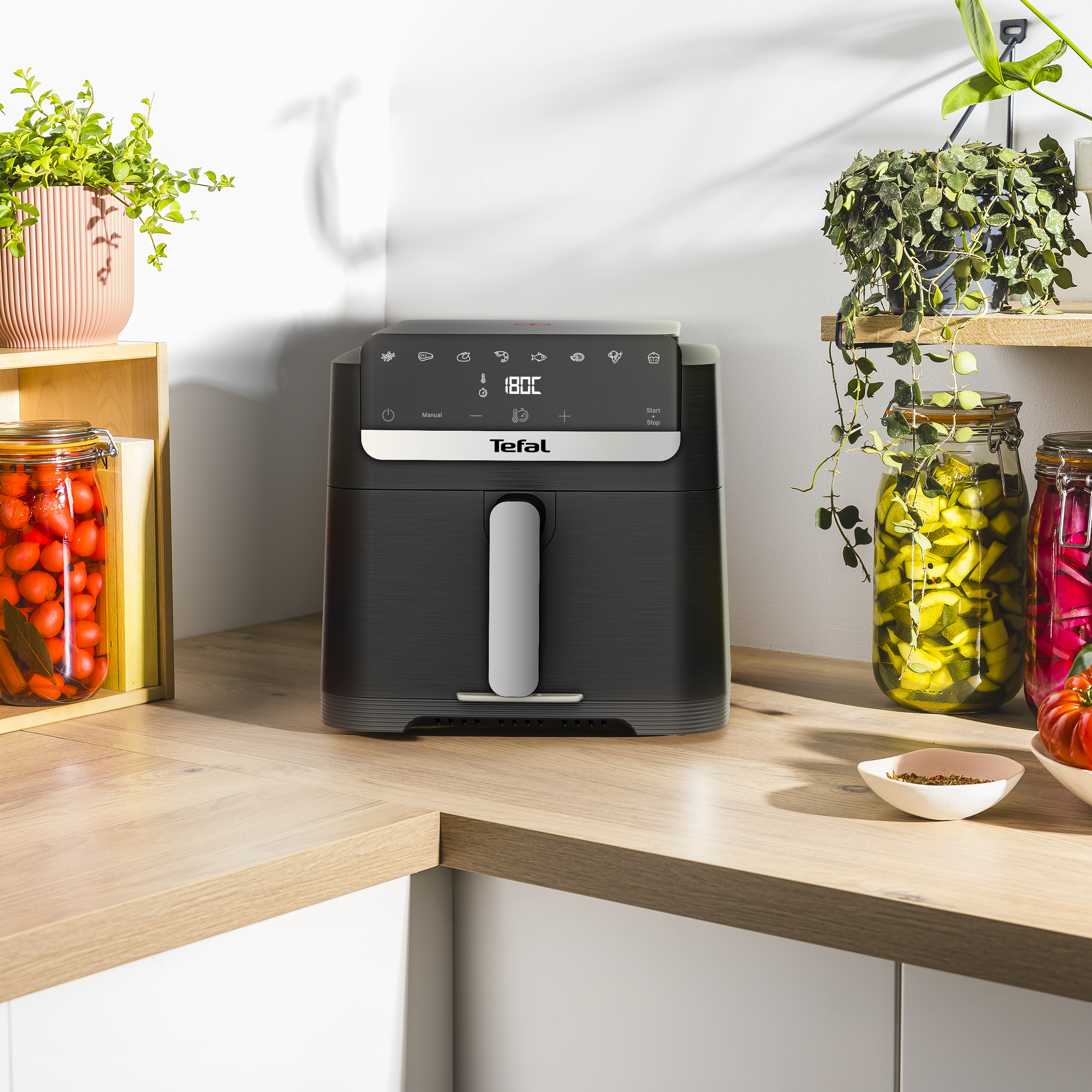 Fekete Tefal légsütő fa munkalapon, befőttesüvegekkel és növényekkel.