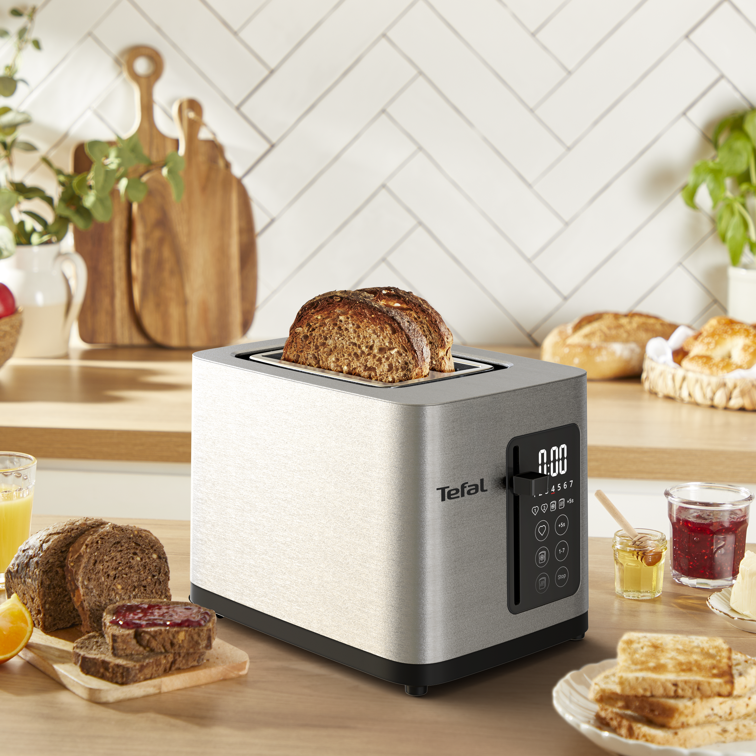 Ezüst színű Tefal kenyérpirító fa asztalon kenyérrel, mézzel, lekvárral és konyhai eszközökkel.