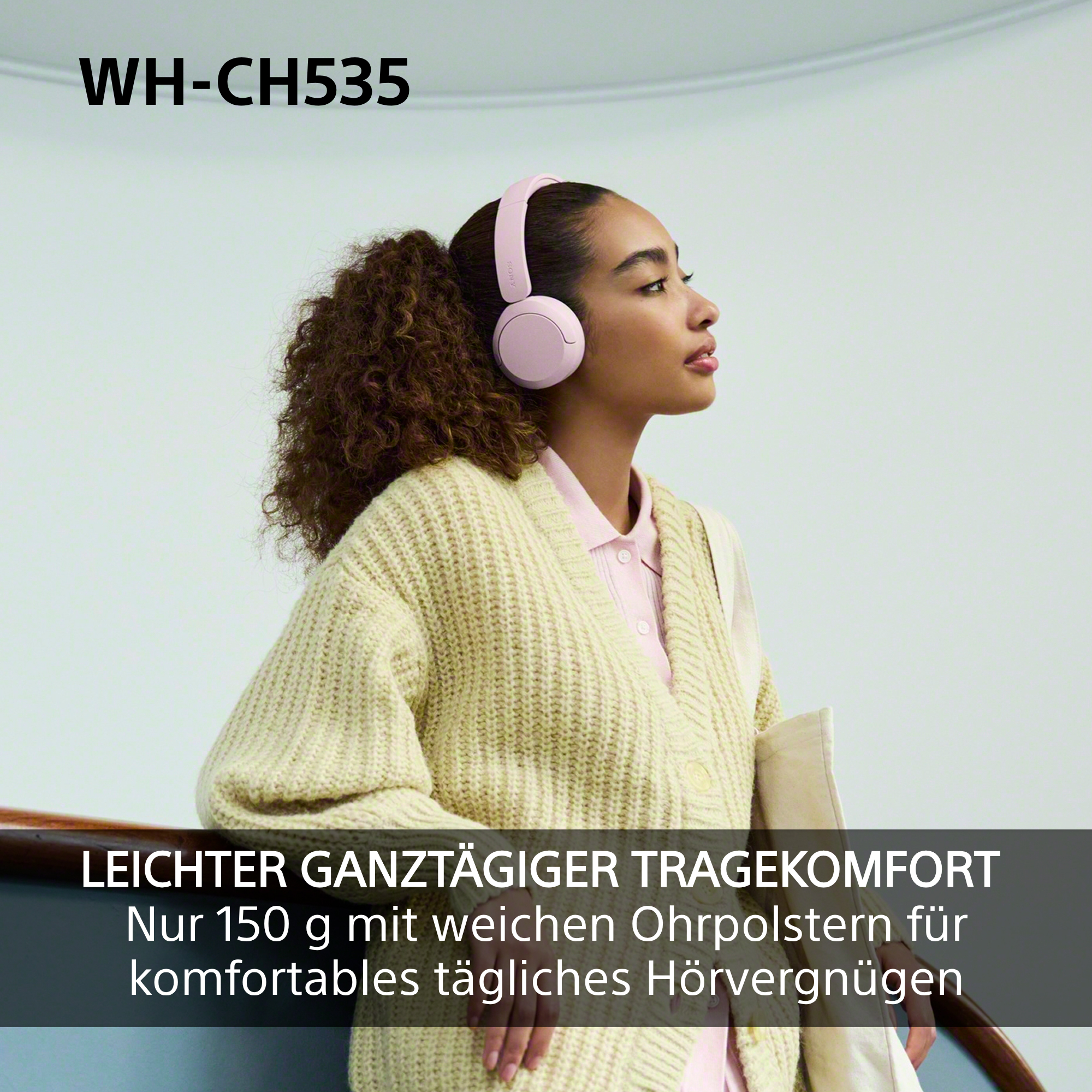 Eine junge Frau mit Locken trägt rosa Kopfhörer und einen beigen Cardigan vor einem hellen Hintergrund.