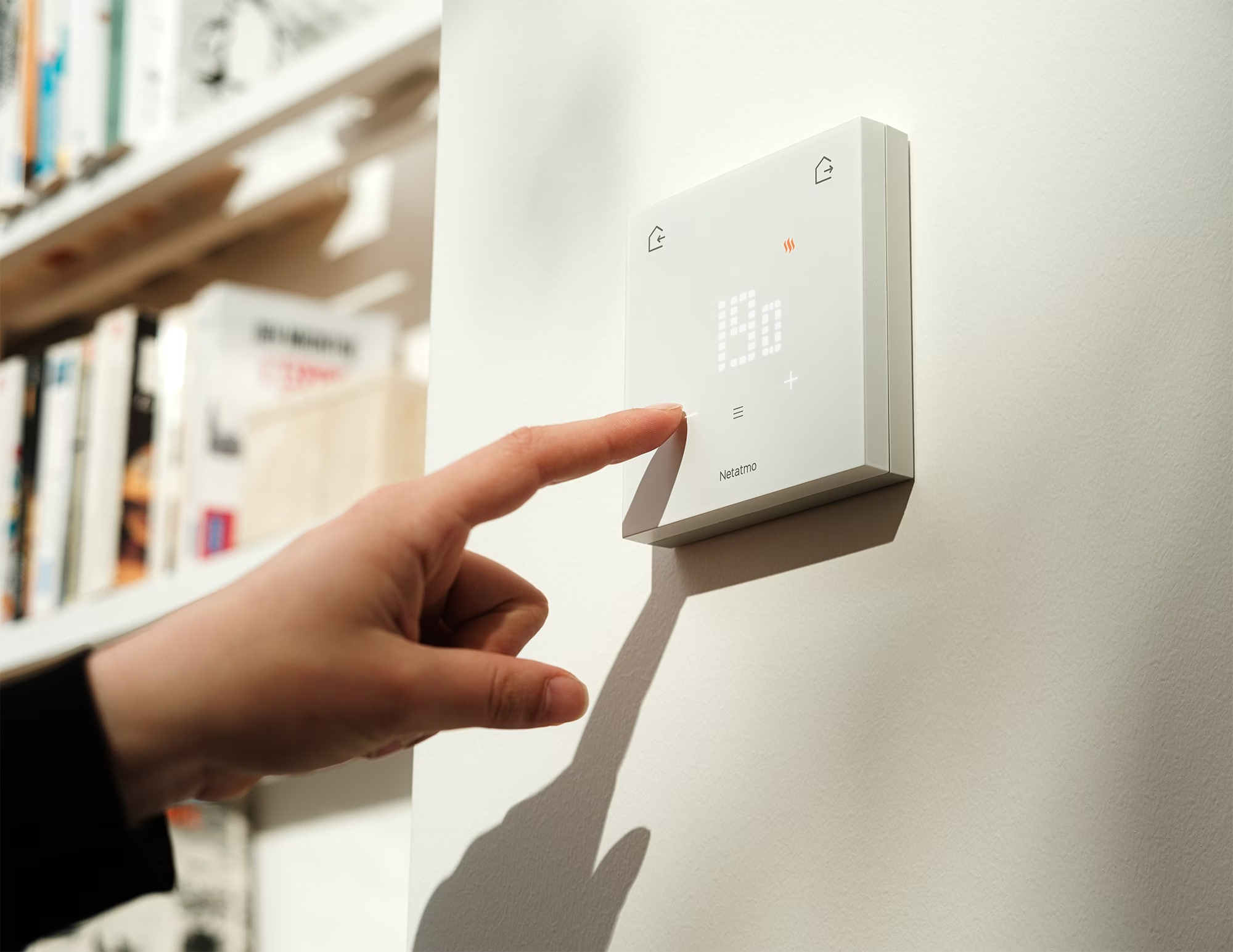 Una mano ajusta un termostato blanco Netatmo en una pared, con una estantería al fondo.