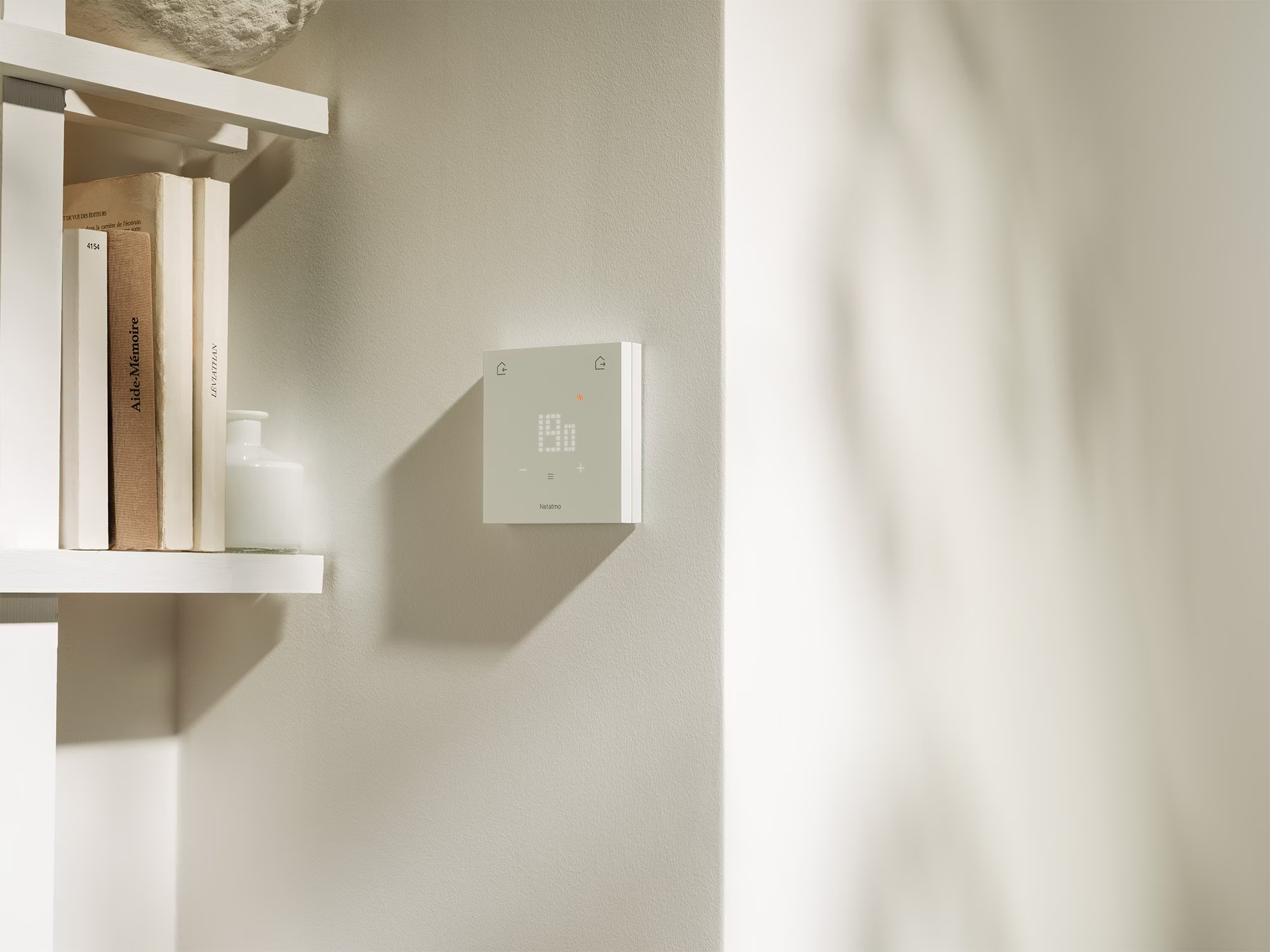 Un termostato Netatmo blanco cuelga de una pared beige junto a una estantería con libros.