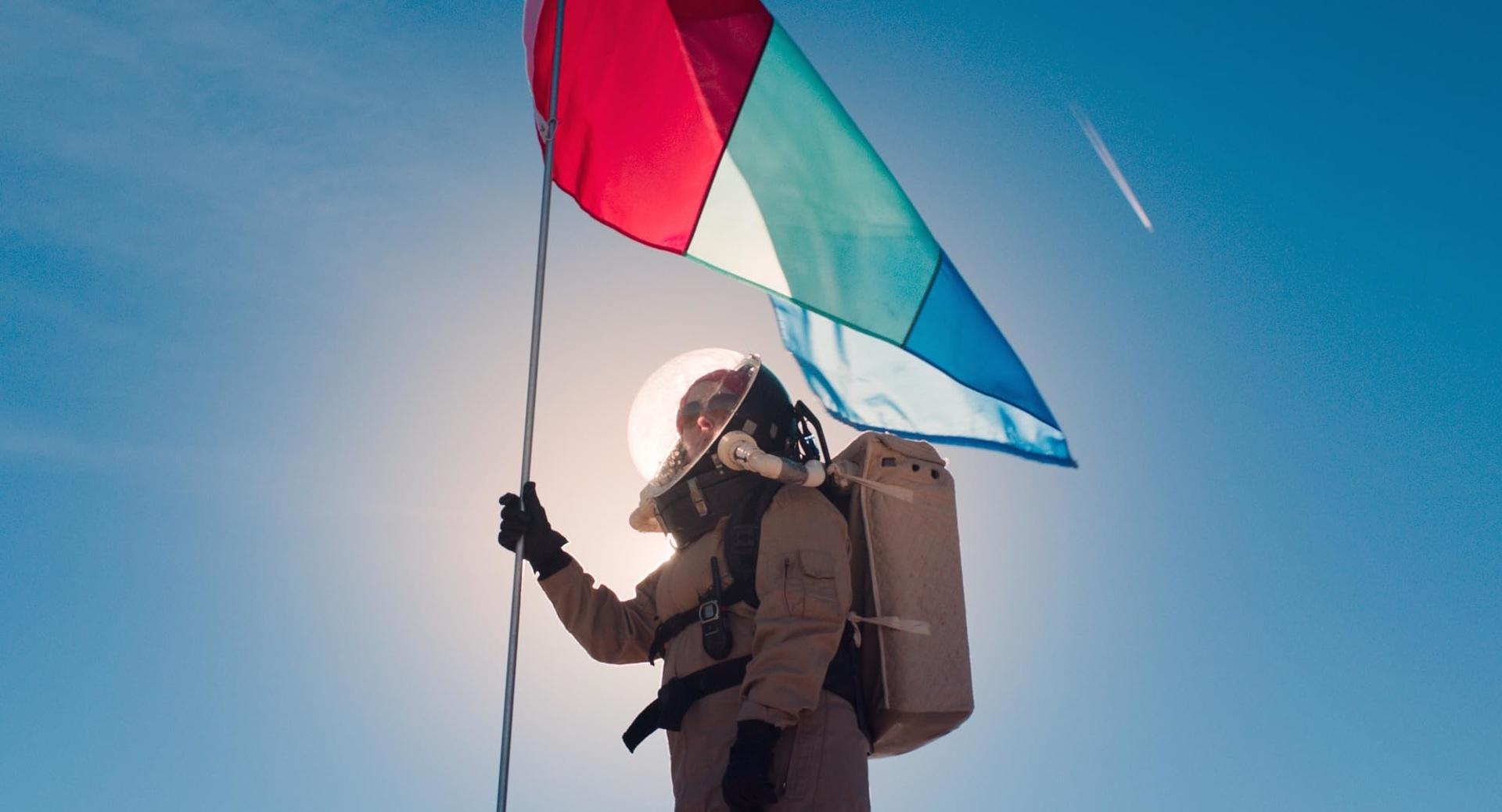 Ein Astronaut im Raumanzug hält eine bunte Flagge vor strahlend blauem Himmel und Sonne.