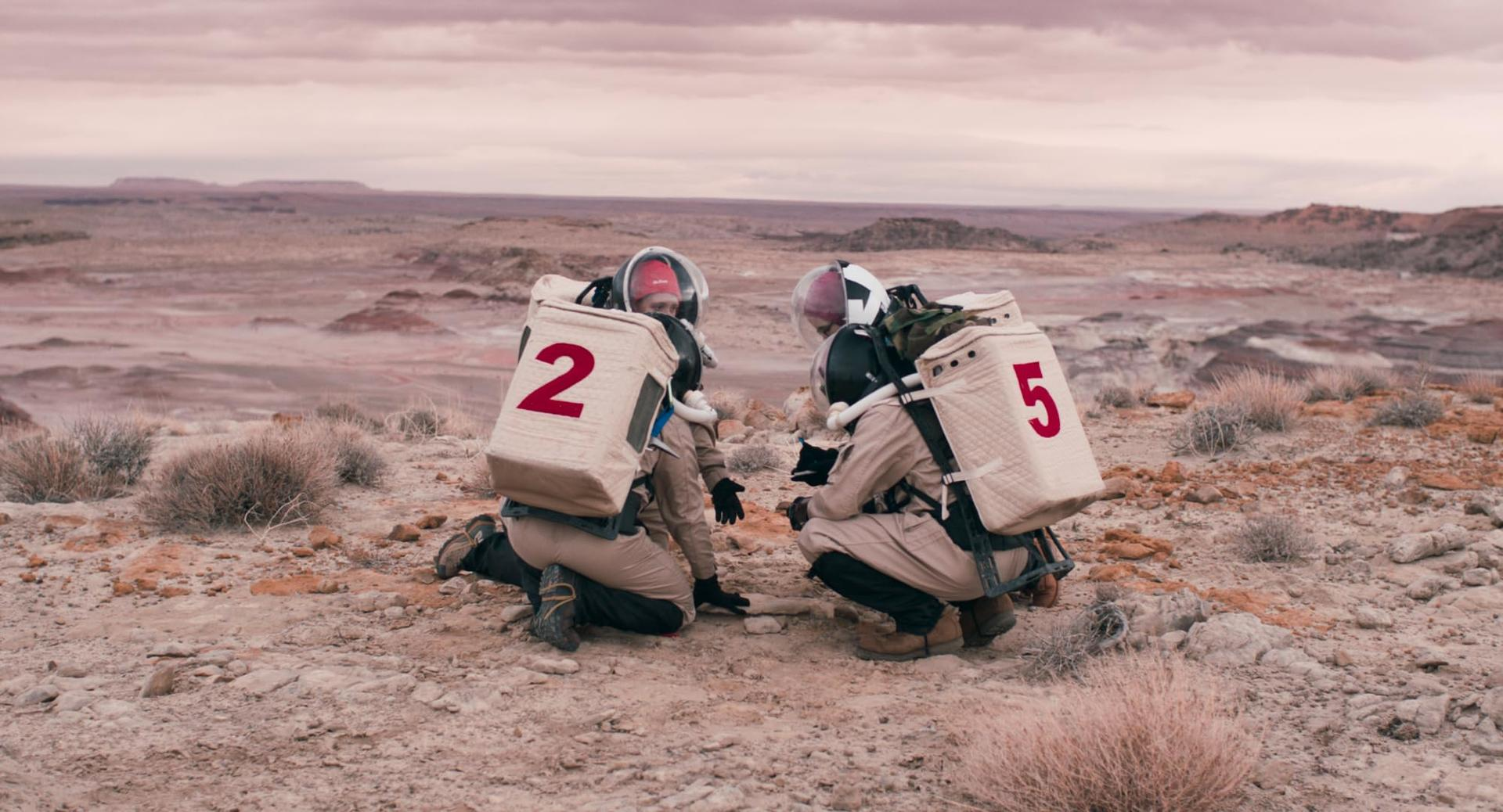 Zwei Astronauten mit den Nummern 2 und 5 hocken in einer kargen, felsigen Landschaft unter einem bewölkten Himmel.
