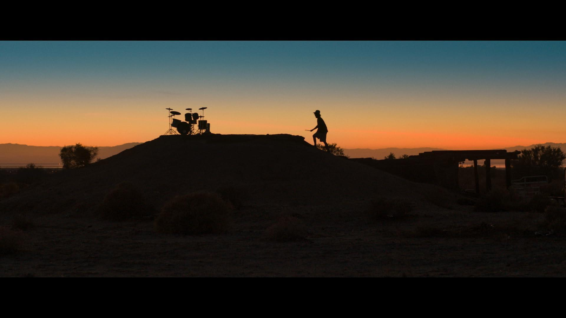 Die Silhouette einer Person und eines Schlagzeugs auf einem Hügel vor einem farbenfrohen Sonnenuntergang.