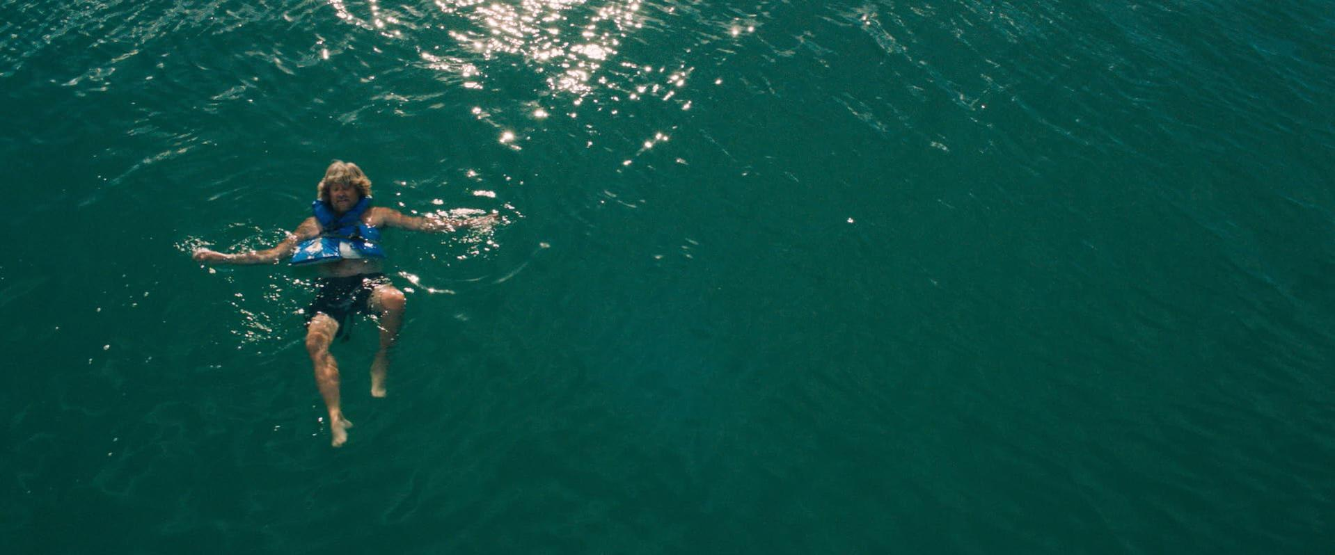 Eine Person mit einer blauen Schwimmweste schwimmt auf dem Rücken im glitzernden, grünen Meerwasser.
