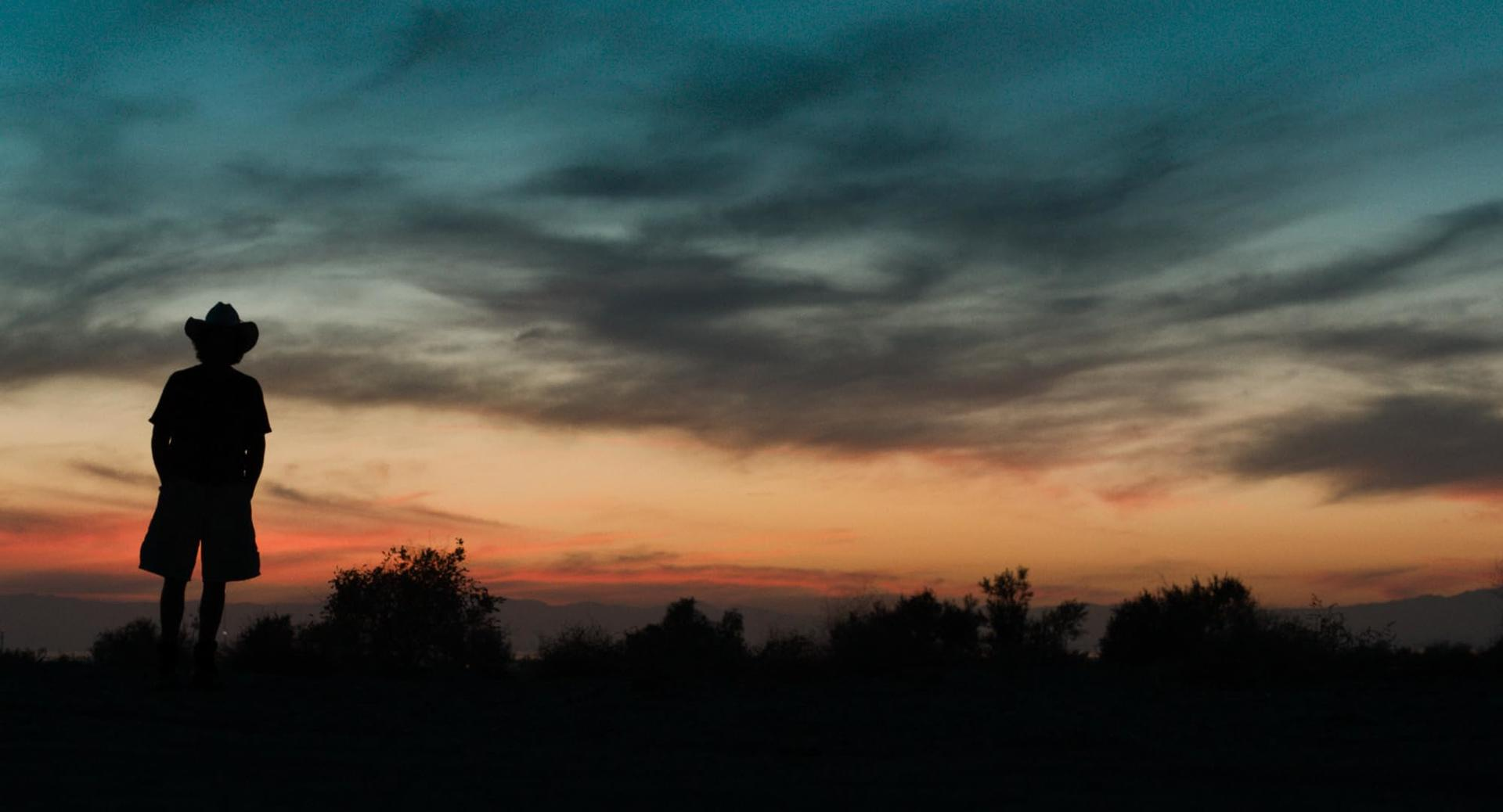 Silhouette einer Person mit Hut vor einem farbenfrohen Sonnenuntergang am Horizont.