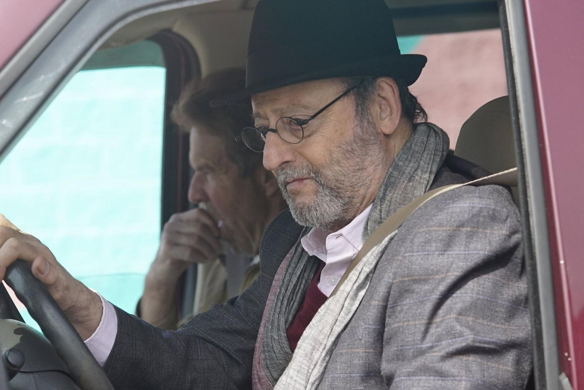 Ein Mann mit schwarzem Hut, Brille und Jacke fährt Auto, ein weiterer Mann sitzt auf dem Beifahrersitz.