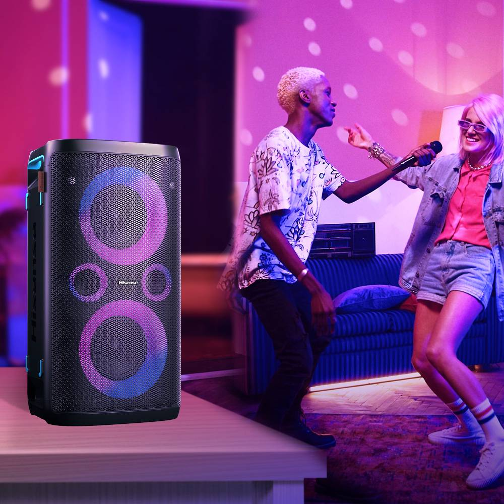 Un gran altavoz portátil con luces en primer plano y dos personas bailando en una habitación iluminada de rosa.