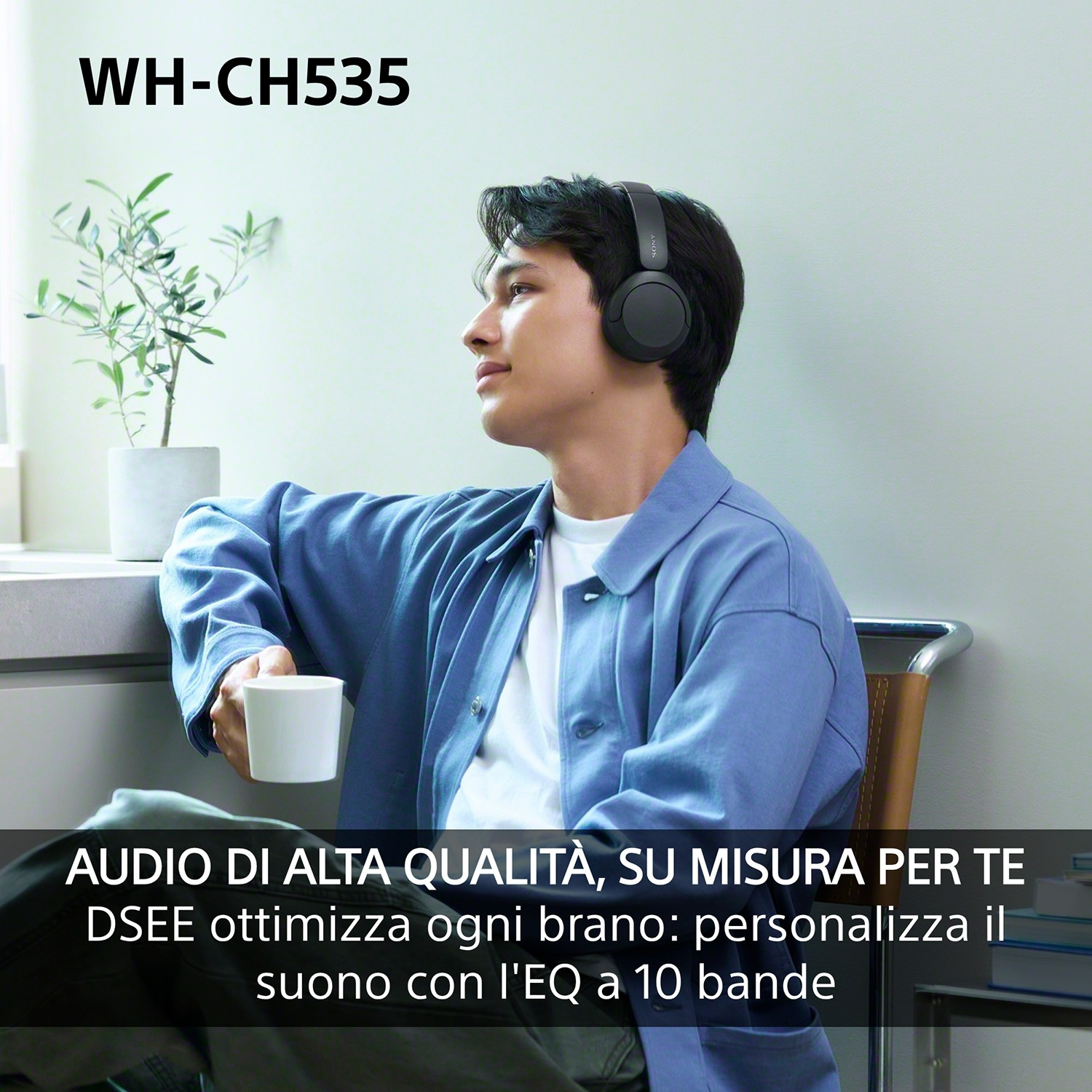 Un giovane con le cuffie tiene in mano una tazza di caffè in una stanza luminosa con una pianta sullo sfondo.