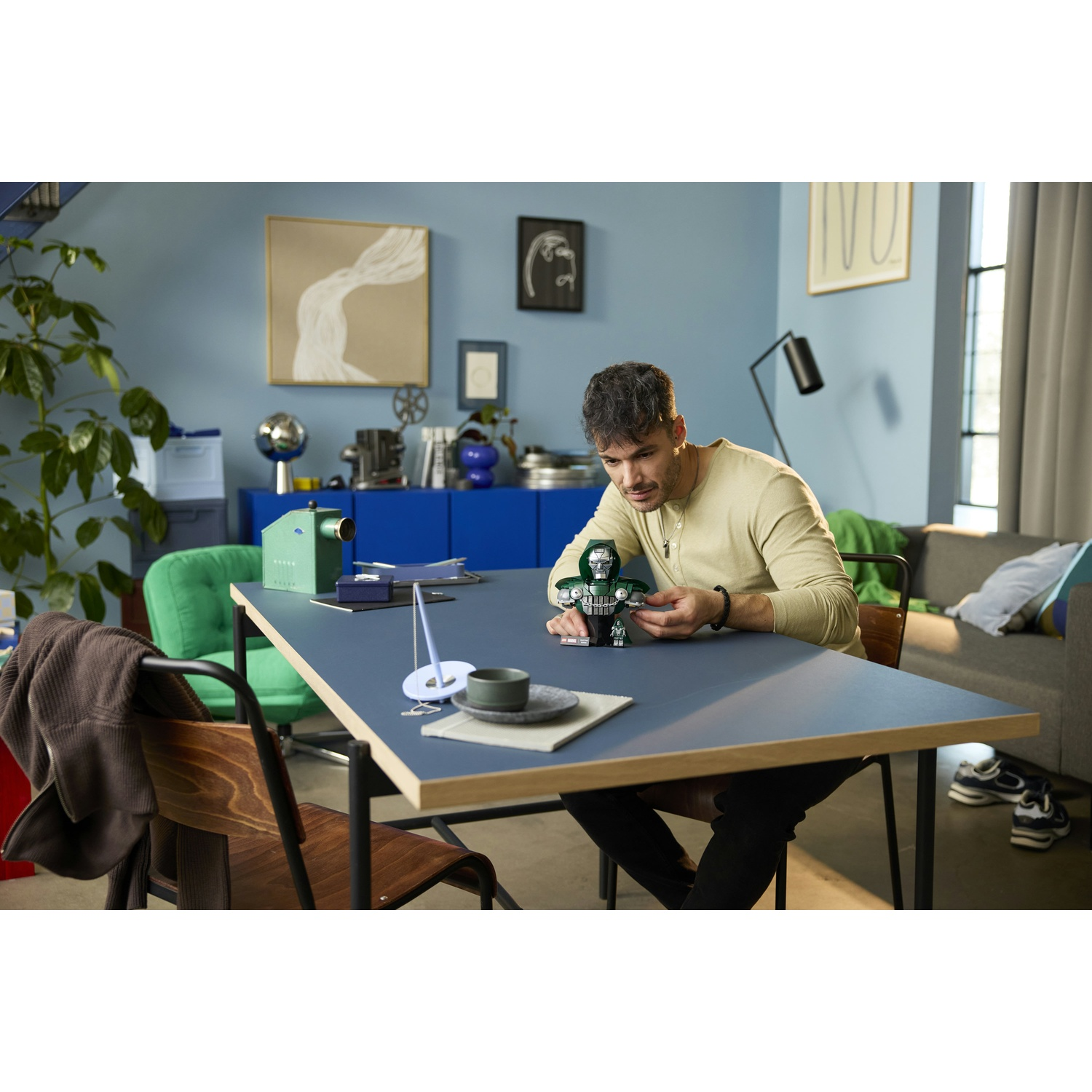Un uomo è seduto a un tavolo blu in una stanza accogliente, mentre assembla un modello Lego verde.