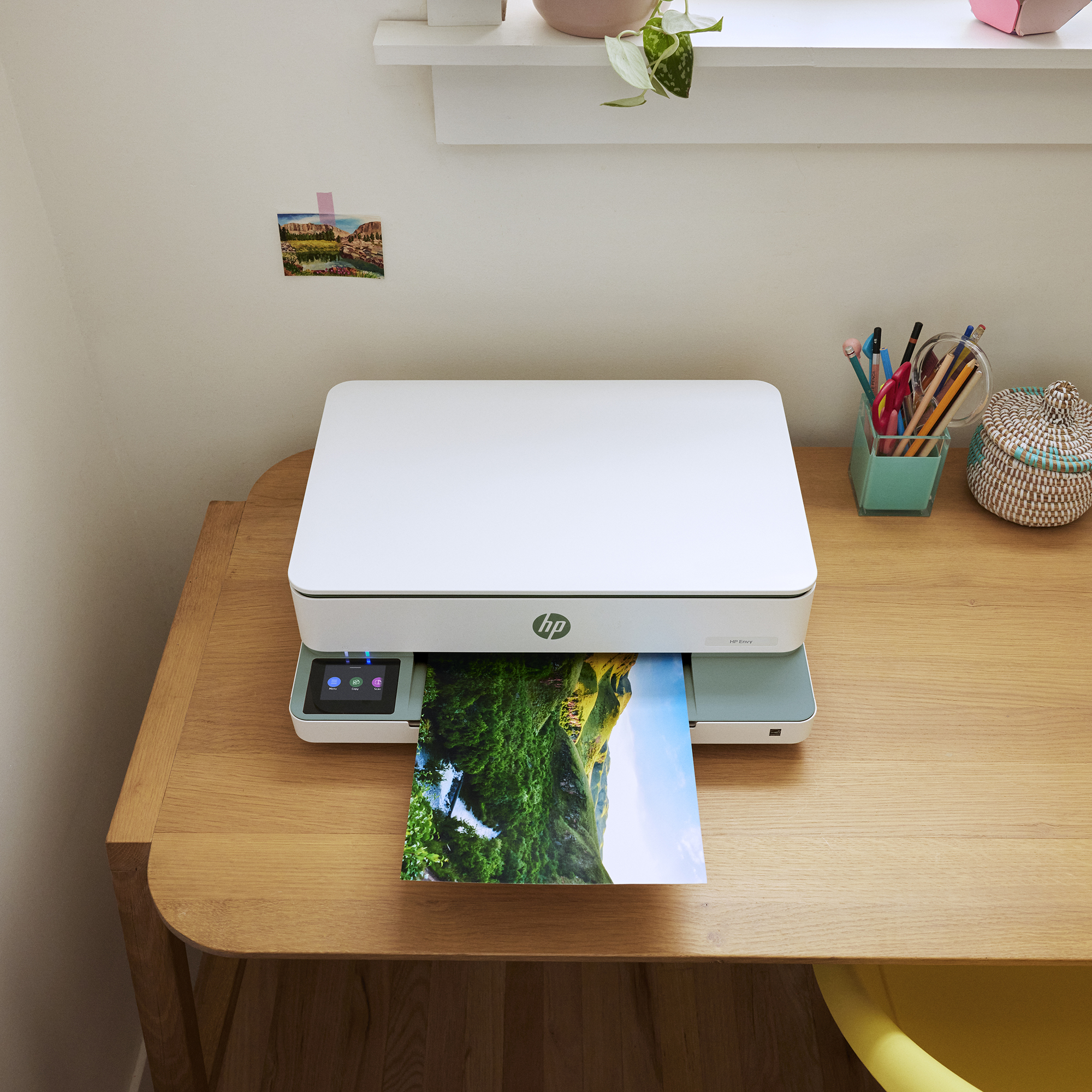 Ein weißer HP Envy Drucker steht auf einem Holztisch und druckt ein farbiges Landschaftsfoto aus.