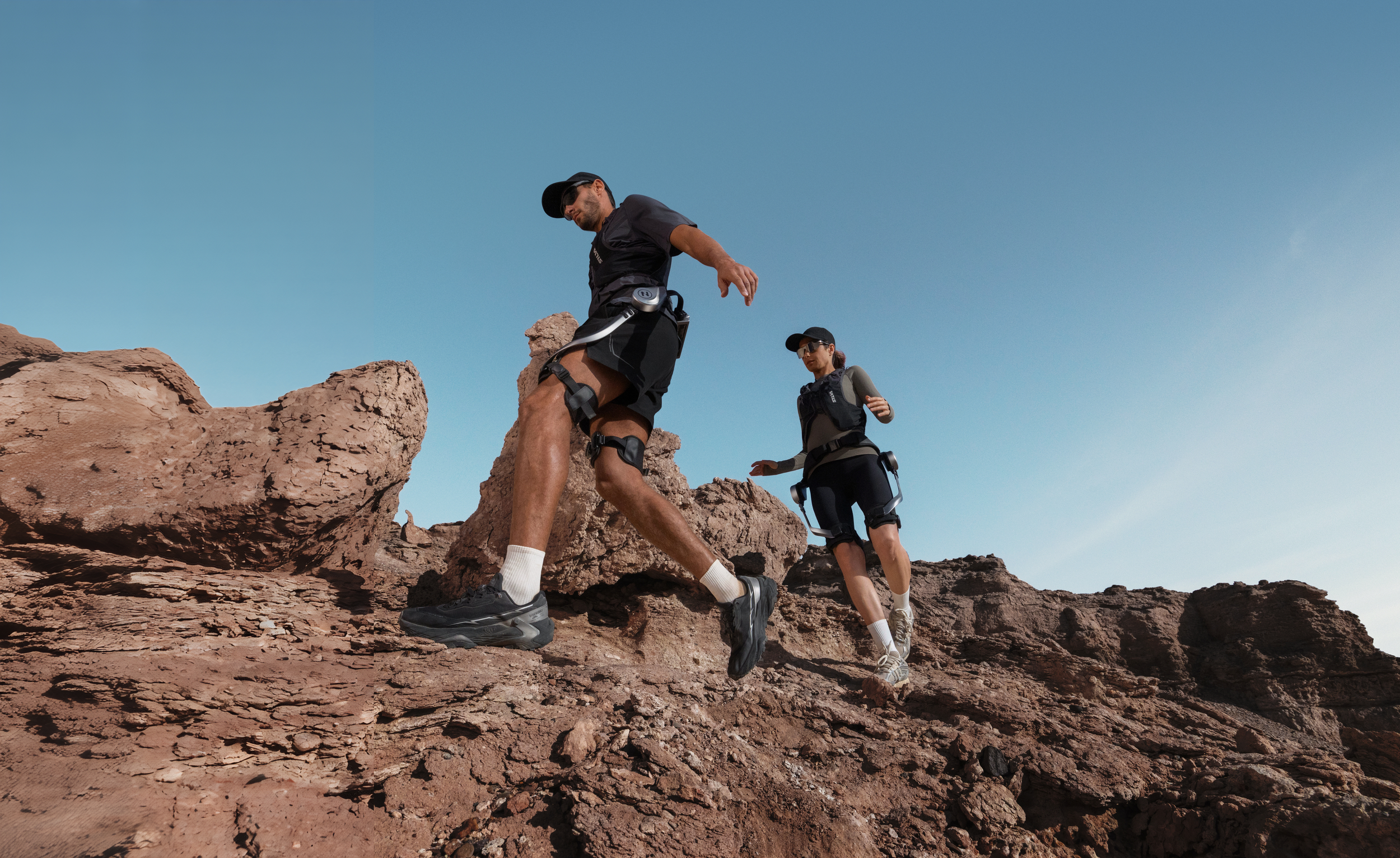 Zwei Personen wandern mit Exoskeletten in einer felsigen Wüstenlandschaft unter blauem Himmel.