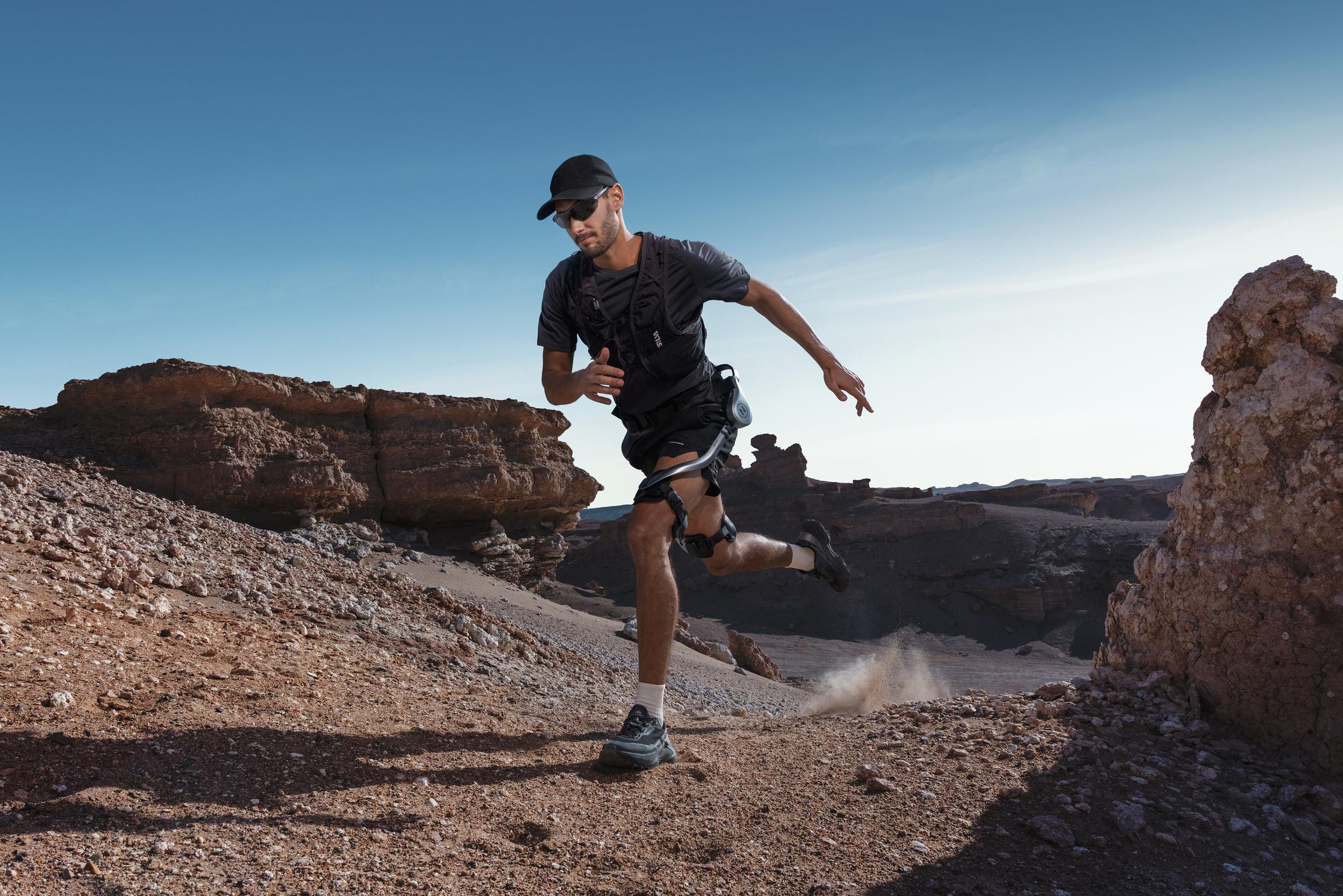 Ein Läufer in schwarzer Sportkleidung und mit einer Exoskelett-Beinhalterung rennt durch eine felsige Wüstenlandschaft.