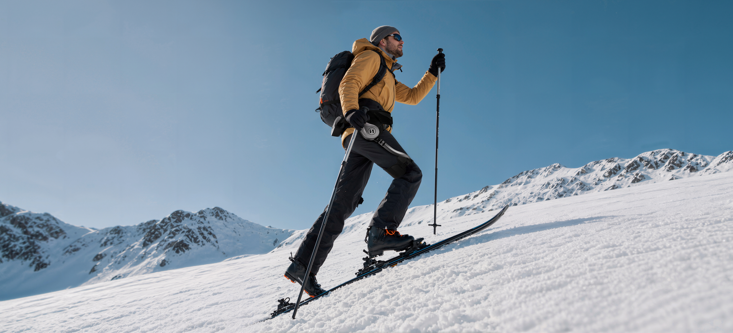 Ein Skifahrer in gelber Jacke besteigt bei klarem blauen Himmel einen schneebedeckten Berghang.