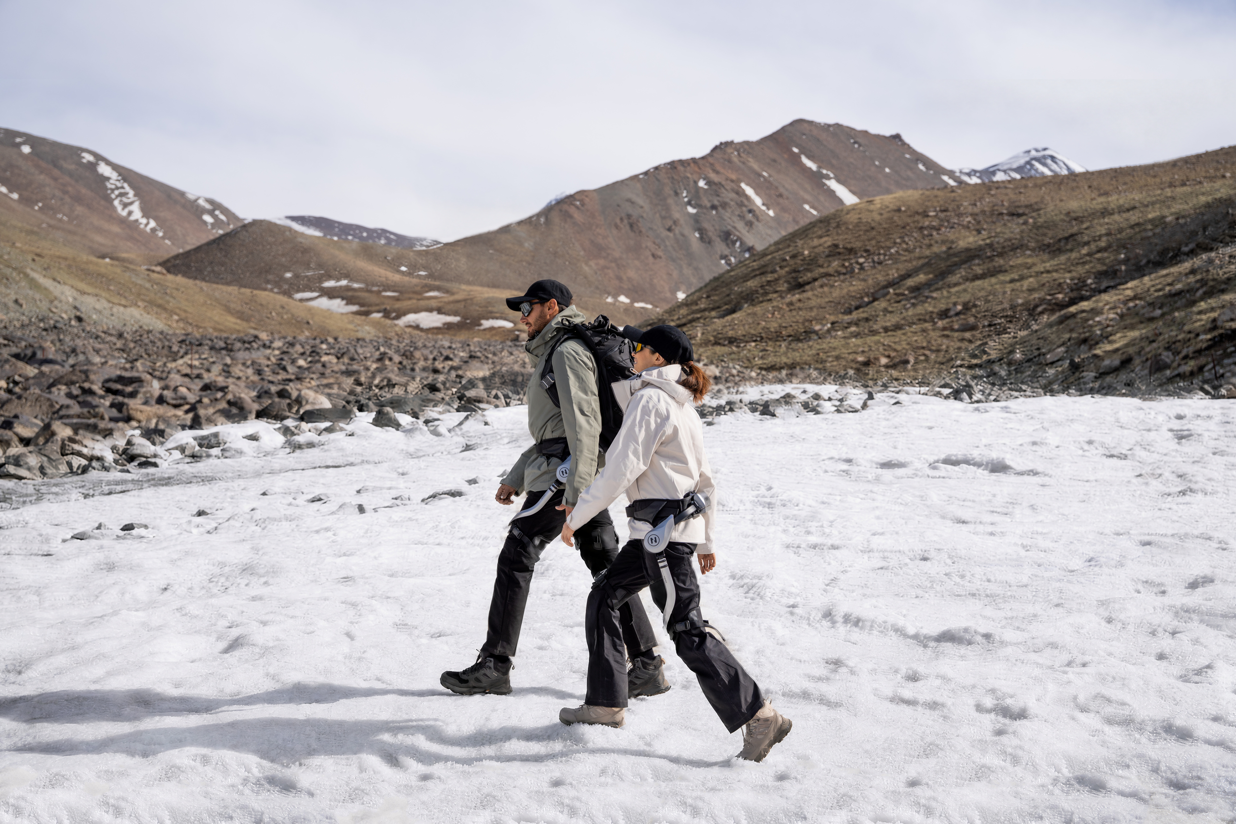 Zwei Wanderer mit Exoskeletten gehen auf einem verschneiten Bergpfad vor einer felsigen Berglandschaft.
