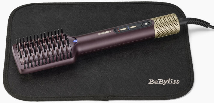 Une brosse lissante BaByliss violette et dorée repose sur un tapis en tissu noir avec le logo BaByliss.
