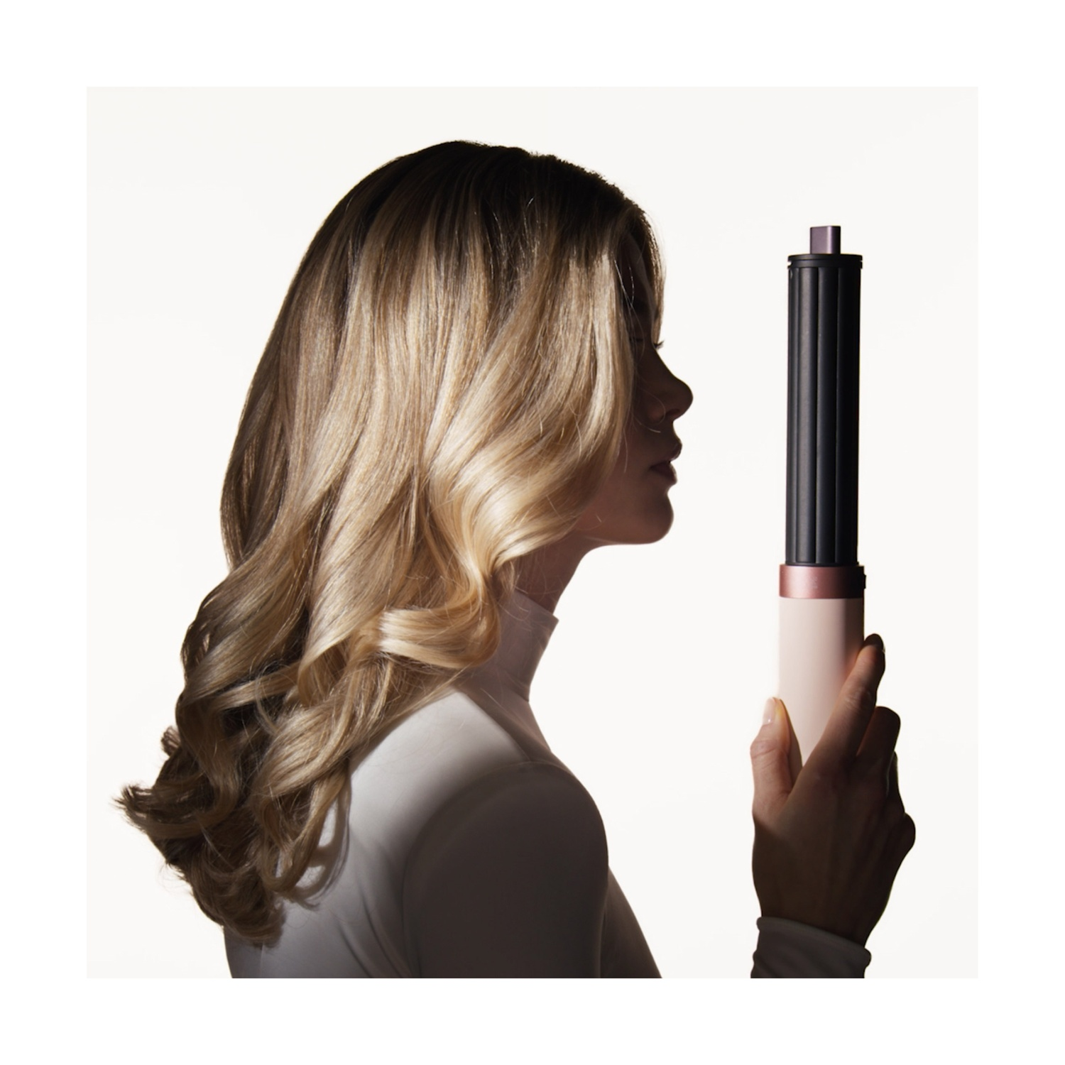 Profilo di una donna bionda con capelli mossi che tiene uno strumento per lo styling su sfondo bianco.