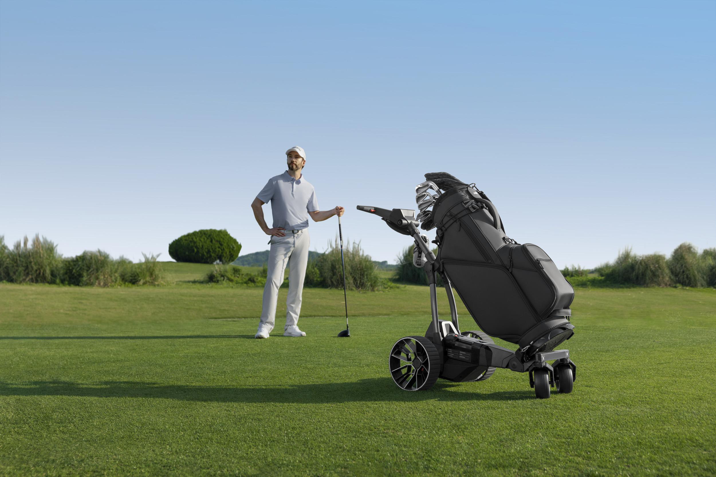 Ein Golfer steht auf einem grünen Golfplatz neben einem motorisierten Golftrolley mit Tasche.